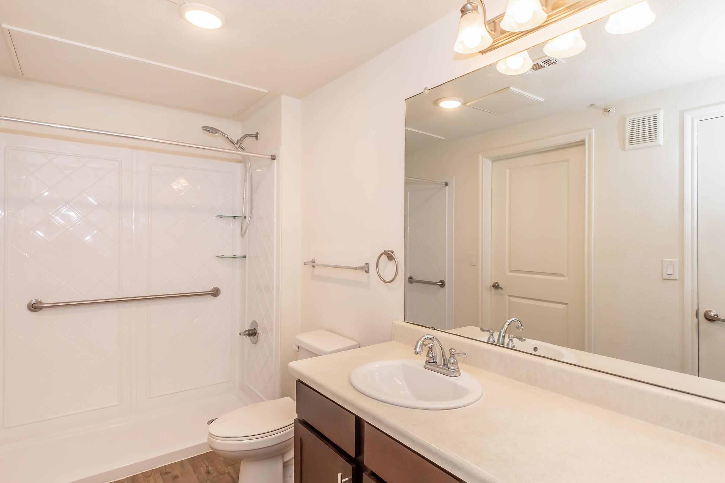 A clean bathroom featuring a white shower with sliding doors, a large mirror above a sink with a faucet, a dark wood vanity with a countertop, a towel rack, and a toilet. Bright overhead lighting and neutral-colored walls create a spacious feel.