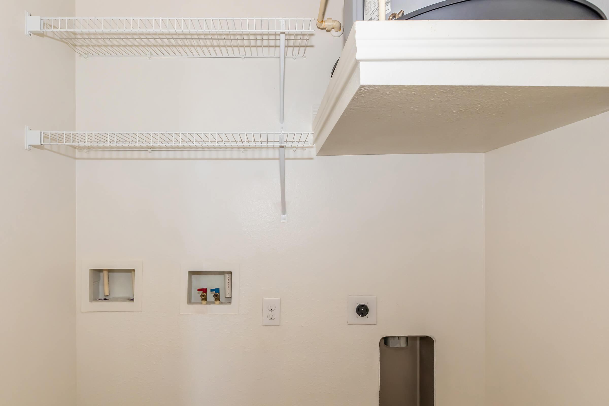 A small, empty laundry room featuring white walls, two wire shelves mounted above, and a small section of wall with two cut-out openings. There is a white utility sink in the corner and plumbing fixtures visible on the wall, creating a functional yet minimalistic space.