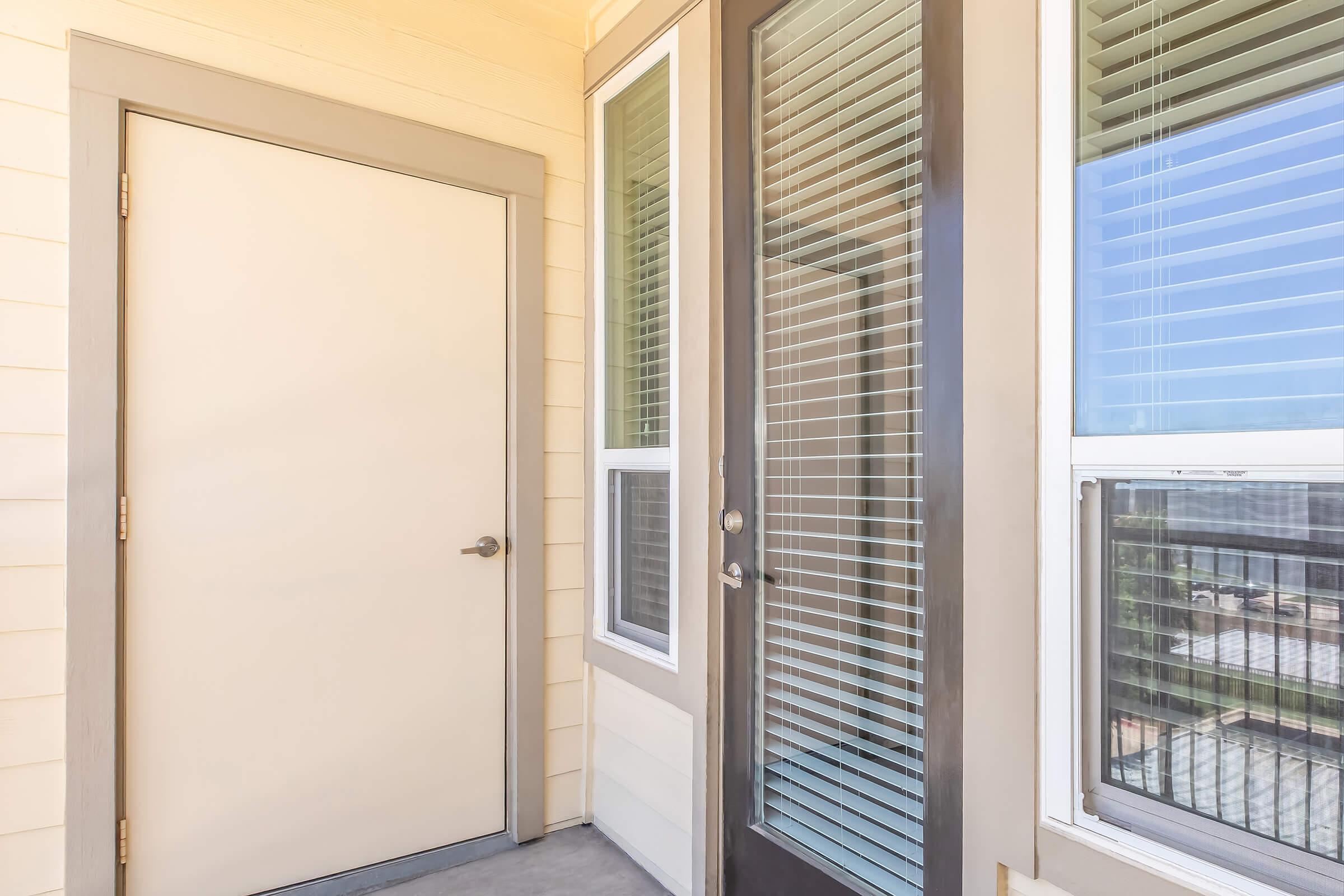 Two doors on a patio: one solid beige door and another with a glass panel and vertical blinds. The exterior features light-colored siding, and a window can be seen beside the glass door, showing a glimpse of the outside view.