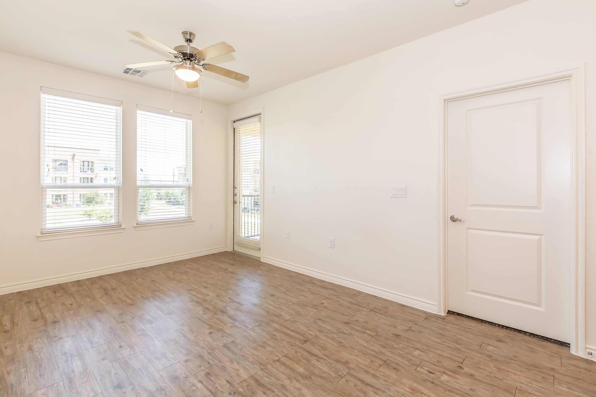 A spacious, empty room featuring light-colored walls, a ceiling fan, and large windows with blinds. The floor is covered with wood-like laminate. There's a door leading to another area, and the overall atmosphere is bright and airy, ideal for personalizing with furniture and decor.