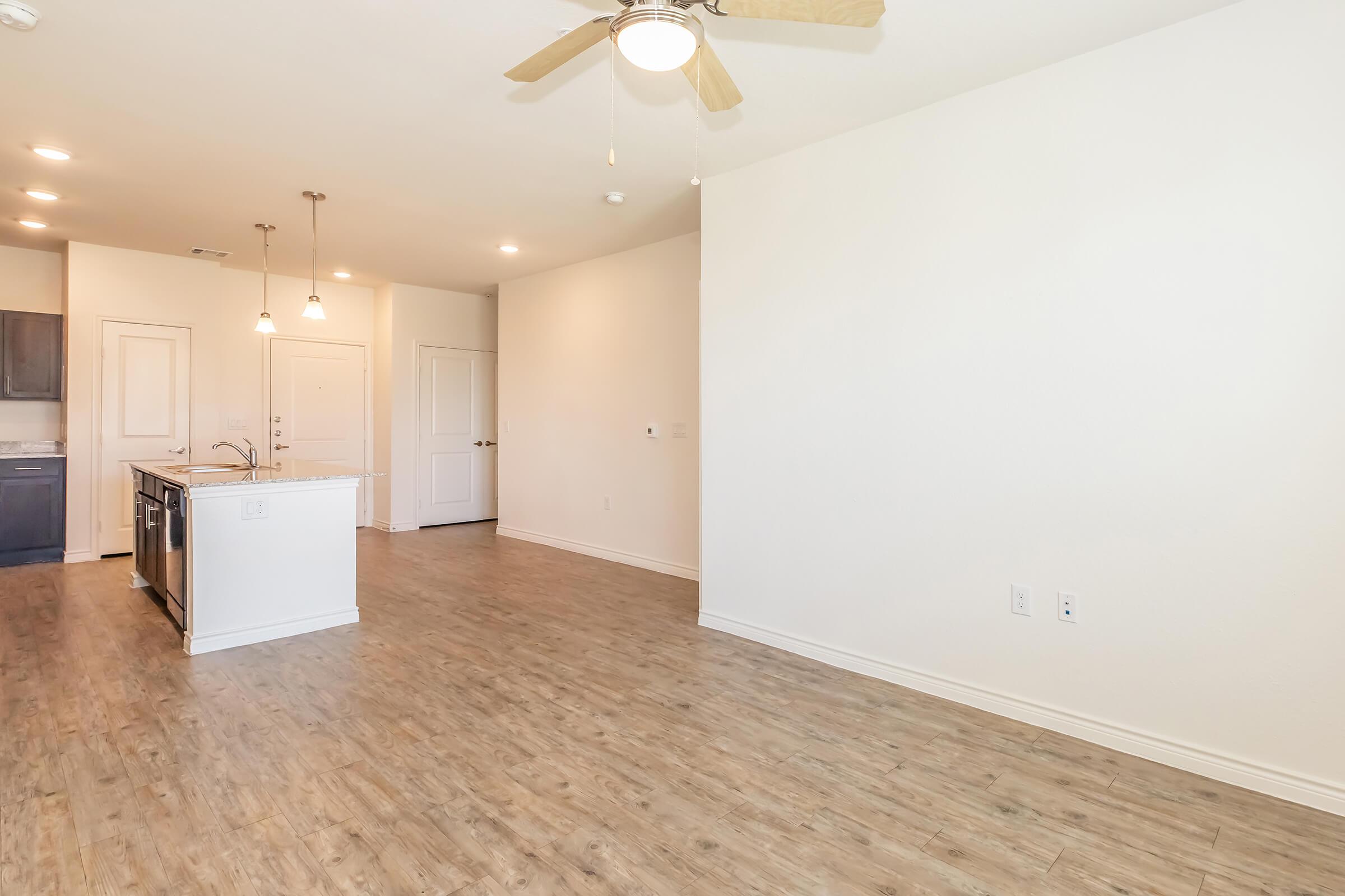 Spacious and bright interior of a modern apartment, featuring light-colored walls, wood-style flooring, and a ceiling fan. The open layout connects a kitchen with a large island to an empty living area, creating a welcoming atmosphere. Natural light fills the space, enhancing the feeling of openness.