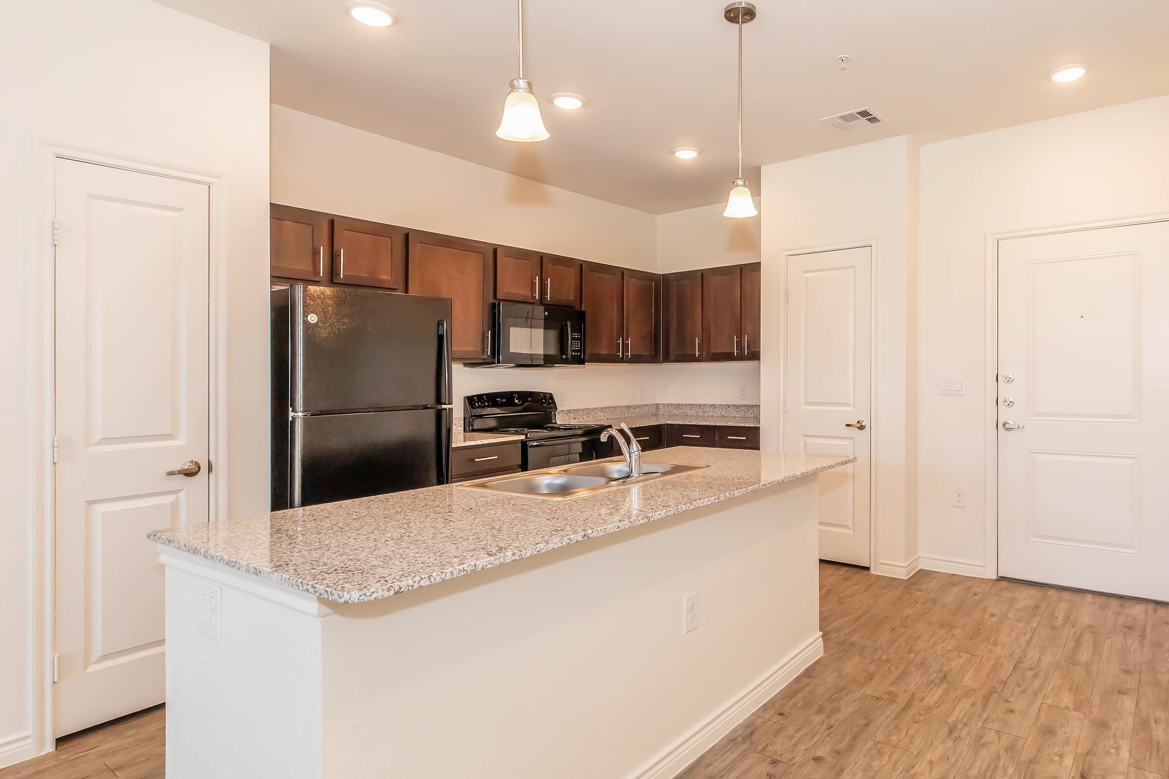 A modern kitchen featuring dark cabinetry, a granite countertop island, stainless steel appliances including a refrigerator and stove, and light fixtures. The space has a warm, inviting atmosphere with wooden flooring and neutral wall colors.