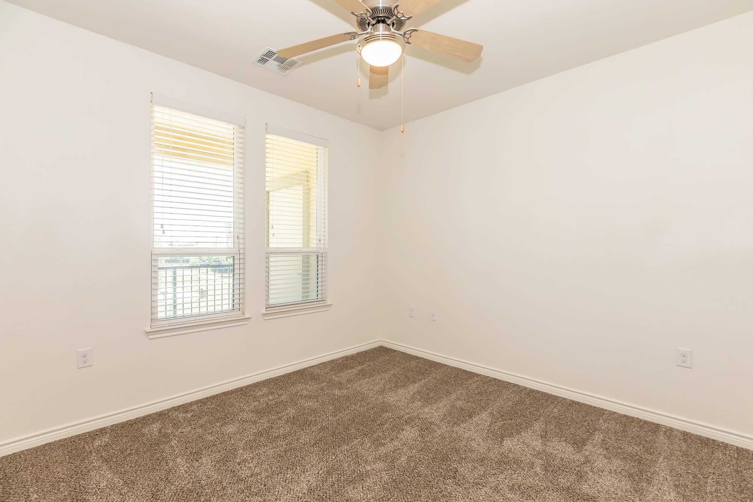 Empty room featuring light-colored walls and carpeted flooring. A ceiling fan is installed, and there are two windows with blinds allowing natural light. The overall atmosphere is bright and spacious.