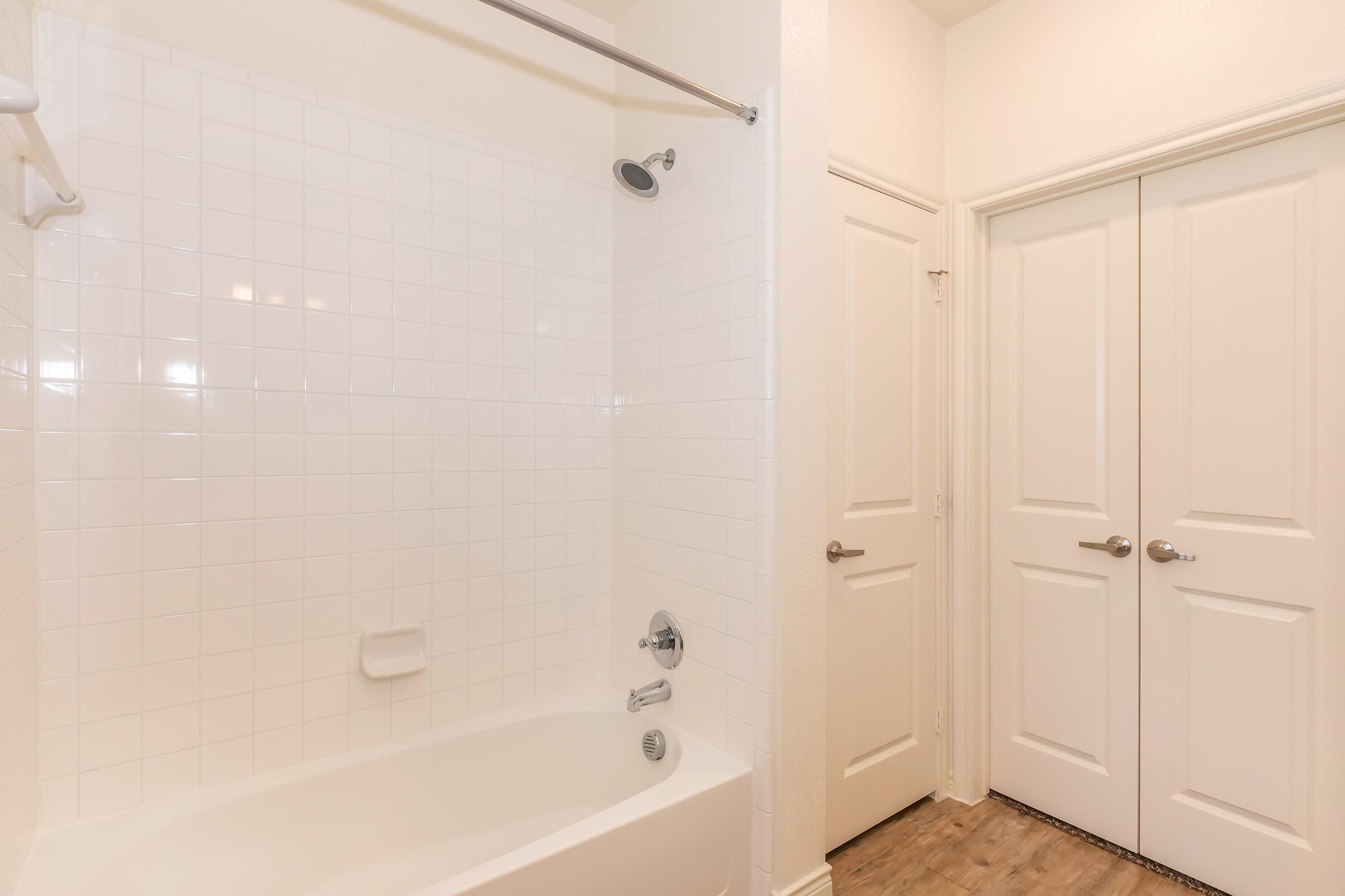 A clean and modern bathroom featuring a white tiled bathtub with a showerhead, and two closed doors in the background, likely leading to a closet or another room. The walls are painted a light color, and the flooring is wooden, contributing to a bright and inviting atmosphere.