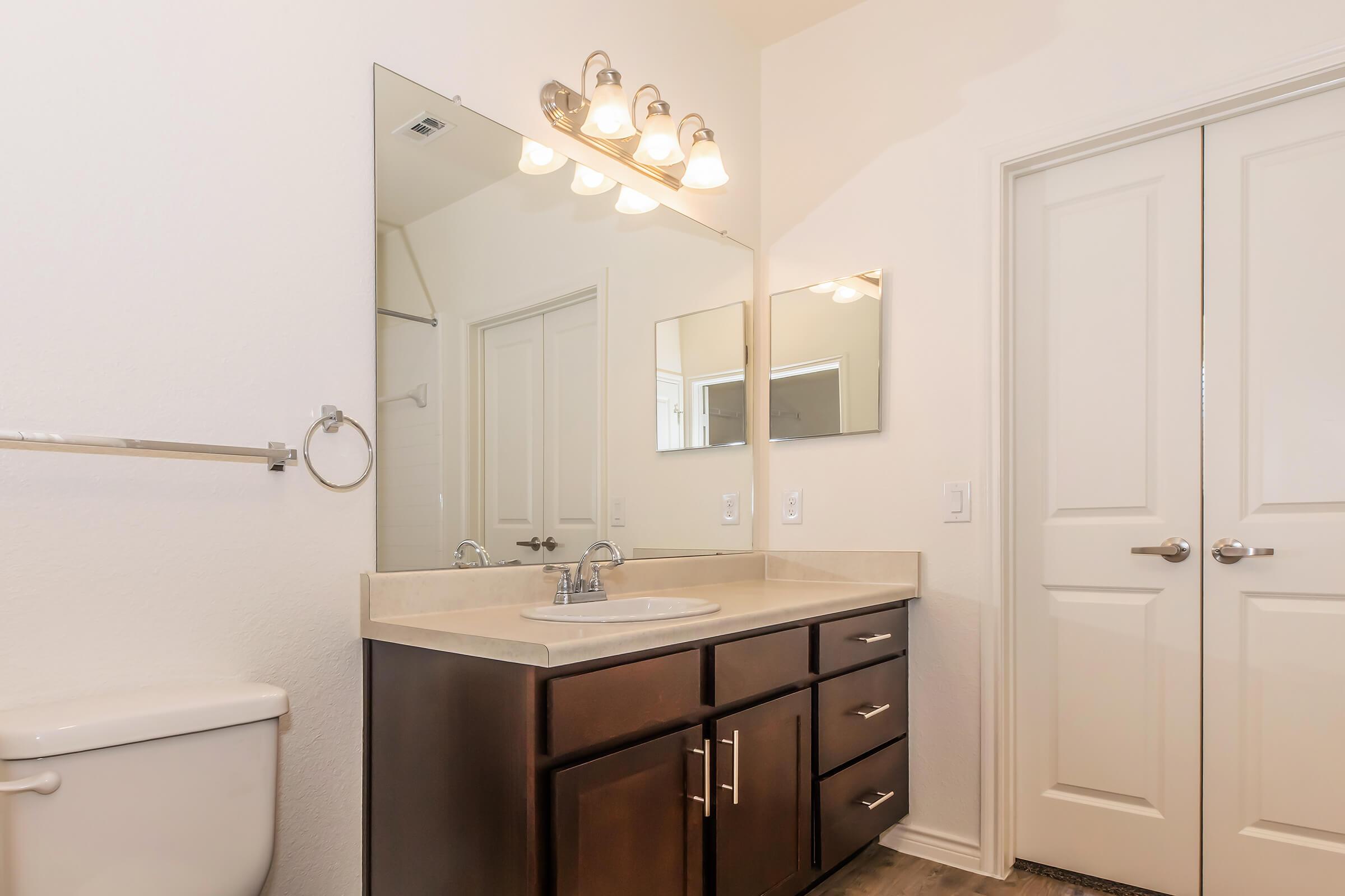 Modern bathroom featuring a double sink vanity with dark wooden cabinets, a large mirror, and three light fixtures above. A white toilet is positioned to the left, and there are two closed doors on the right, indicating closet or storage space. The walls are painted in a soft, neutral color.