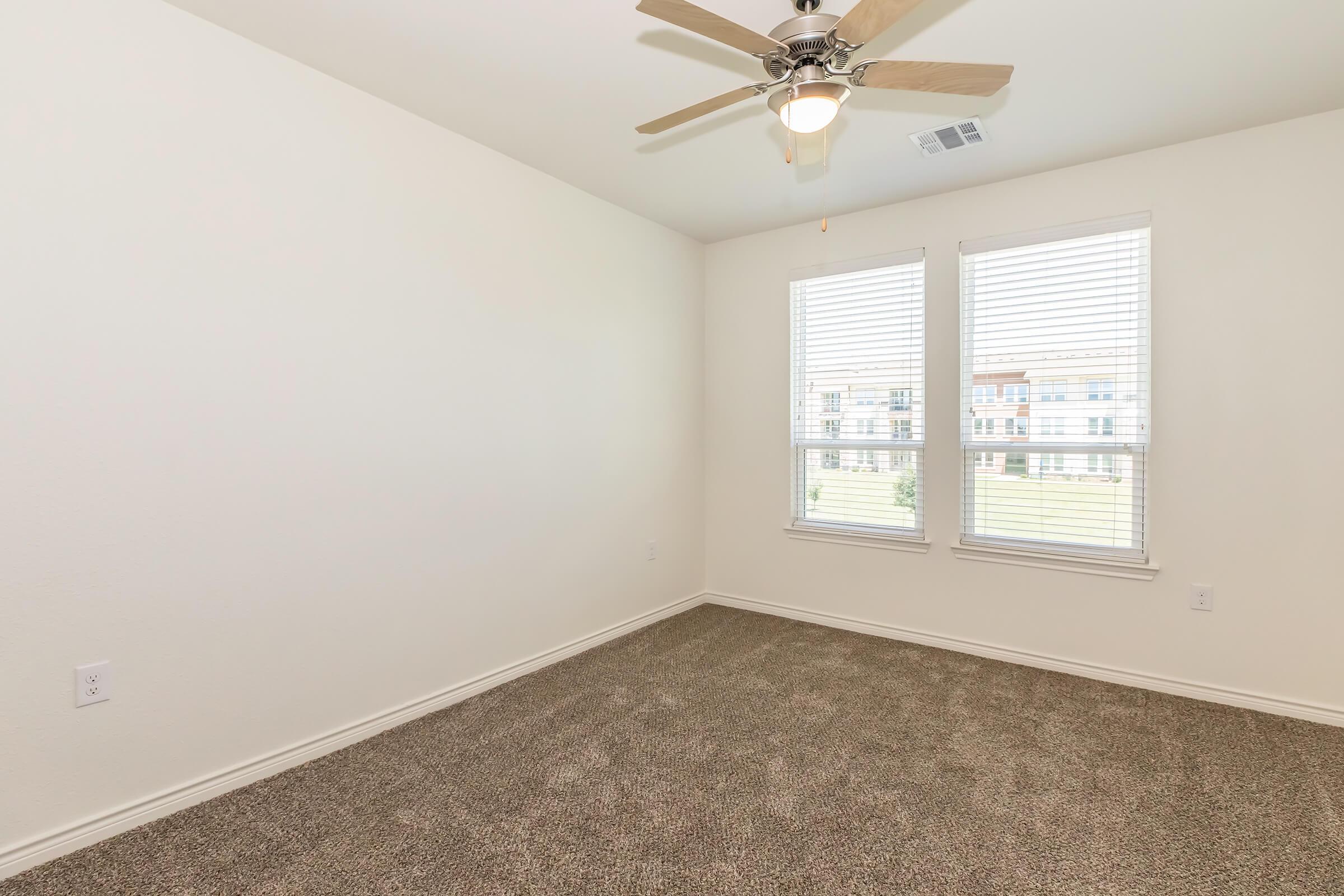 A well-lit, empty room featuring light-colored walls and carpet. It has a ceiling fan and two windows with blinds, allowing natural light to enter. The room has a clean and spacious feel, suitable for various uses such as a bedroom or office. The view outside includes grassy areas and buildings in the background.