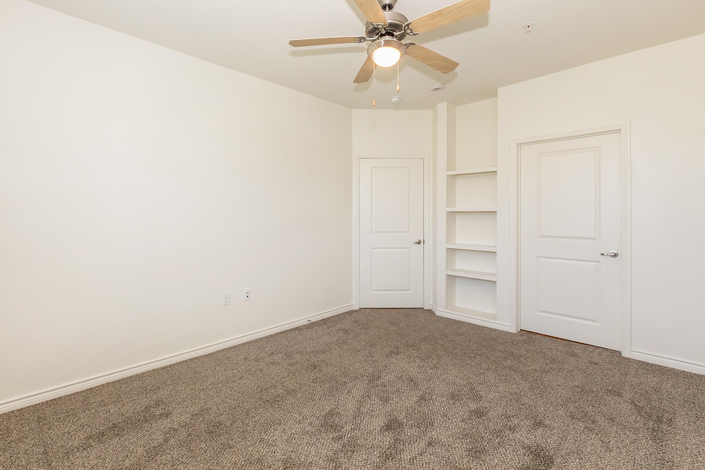 Empty room with beige carpet, a ceiling fan with wooden blades, and white walls. A set of double doors and a built-in shelf are visible on one side, while the opposite wall is bare, creating a spacious and simple atmosphere suitable for various uses.