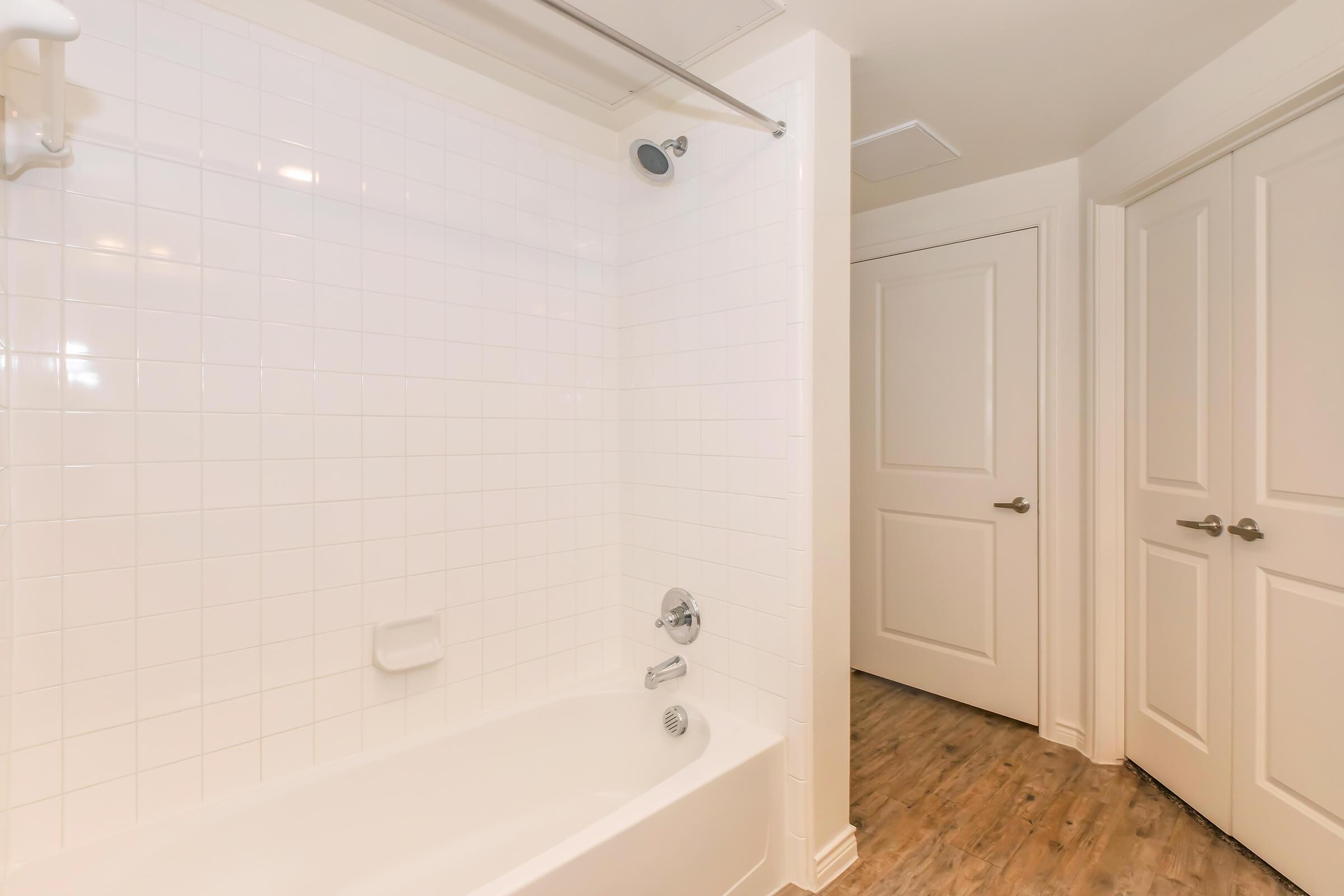 A clean, modern bathroom featuring a white tiled bathtub and shower area with a silver showerhead. The room has a light, neutral color palette, and a door leading to another space is visible in the background. The floor is made of light wood, adding warmth to the design.