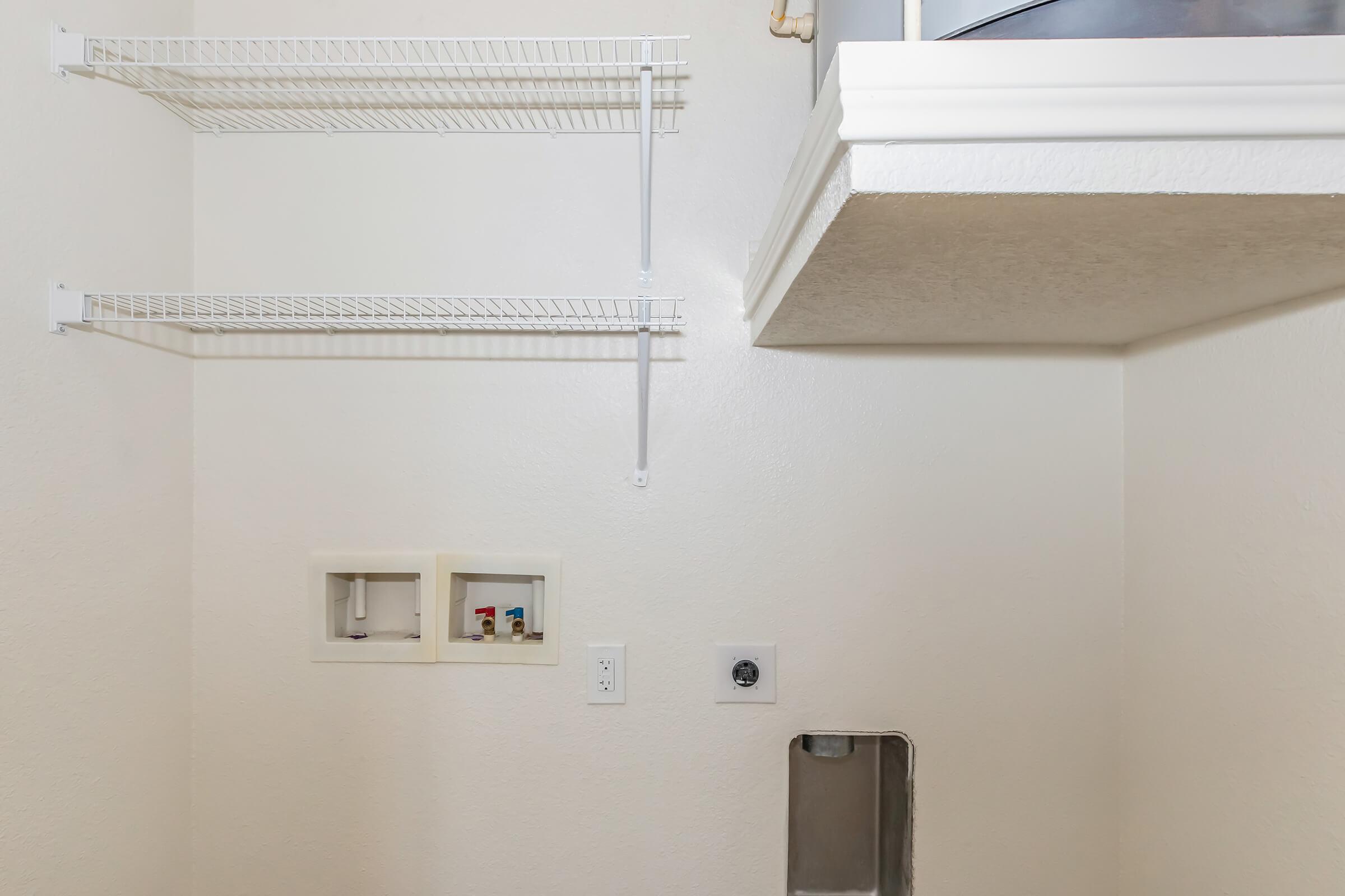 A clean, empty laundry room with white walls. There are two shelving units: one wire shelf mounted on the wall and a small ledge above. A small cabinet inset into the wall holds various utilities. An outlet and water connection are visible on the wall, indicating laundry capabilities.