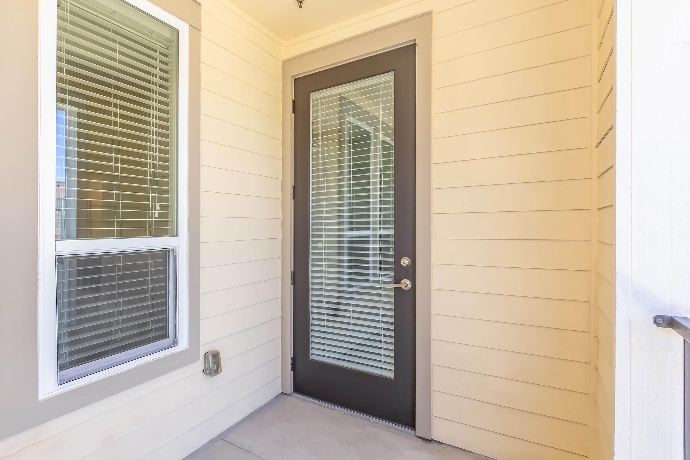 A view of a modern entrance featuring a single door with glass panels, framed by beige exterior walls and a window to the left, allowing natural light to enter the space. The floor is concrete, giving a clean and minimalistic look.