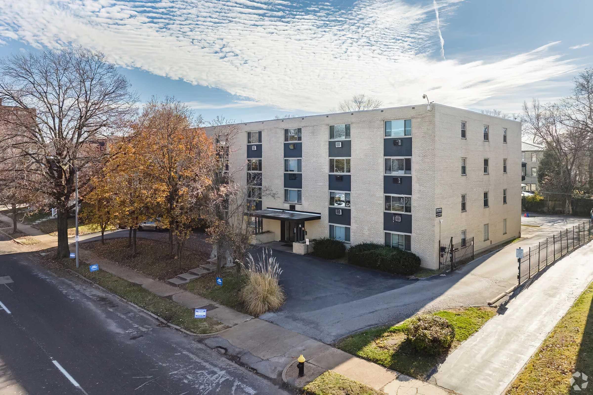 A four-story apartment building made of light-colored brick, surrounded by a few trees. The facade features multiple windows and a covered entrance. The scene is set on a clear day with scattered clouds in the background, and the street is lined with parked cars and signs.