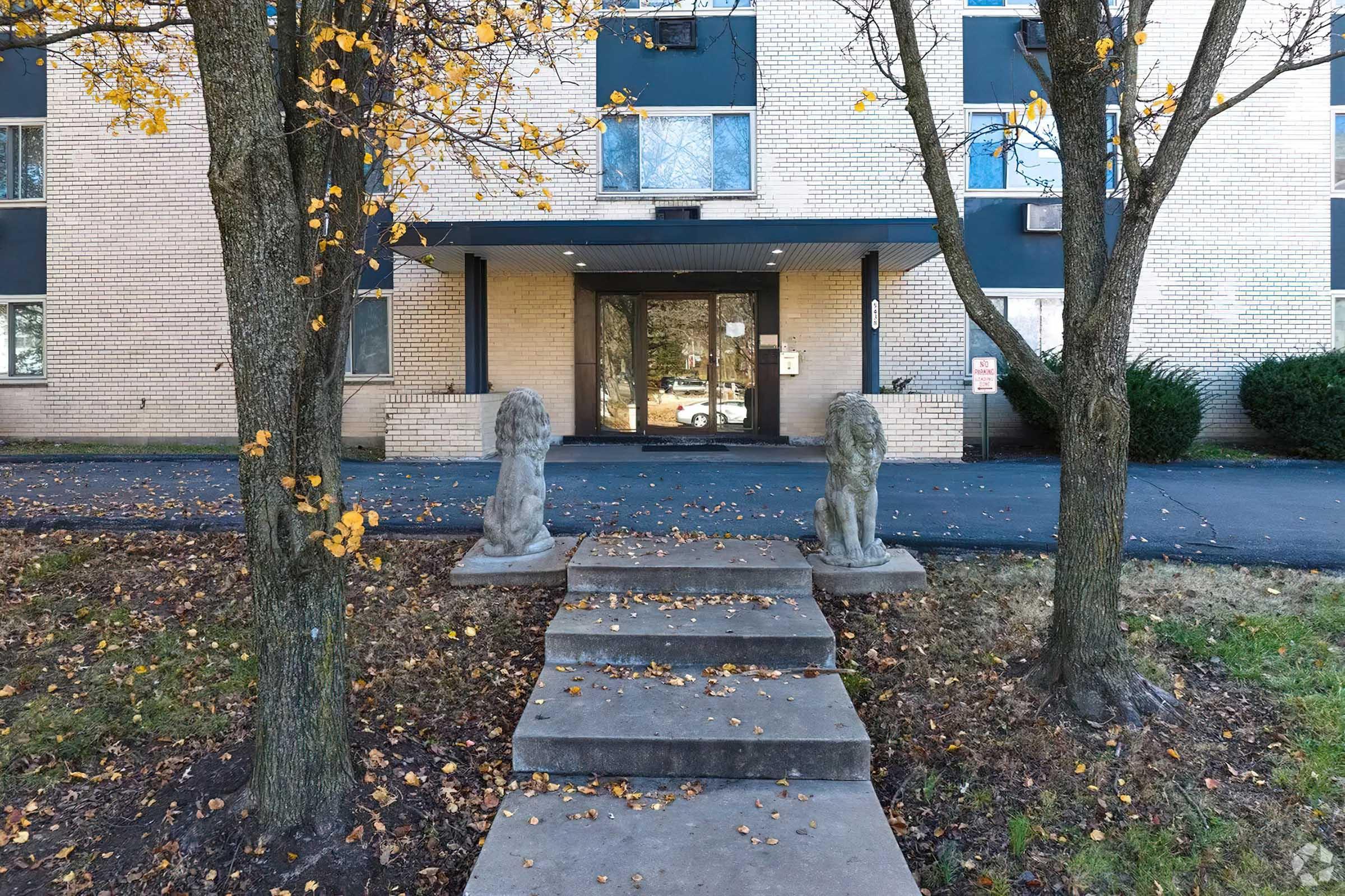 A pathway leads to the entrance of a building, flanked by two stone lion statues. Trees with autumn leaves border the walkway, and the building features a brick facade and large windows.
