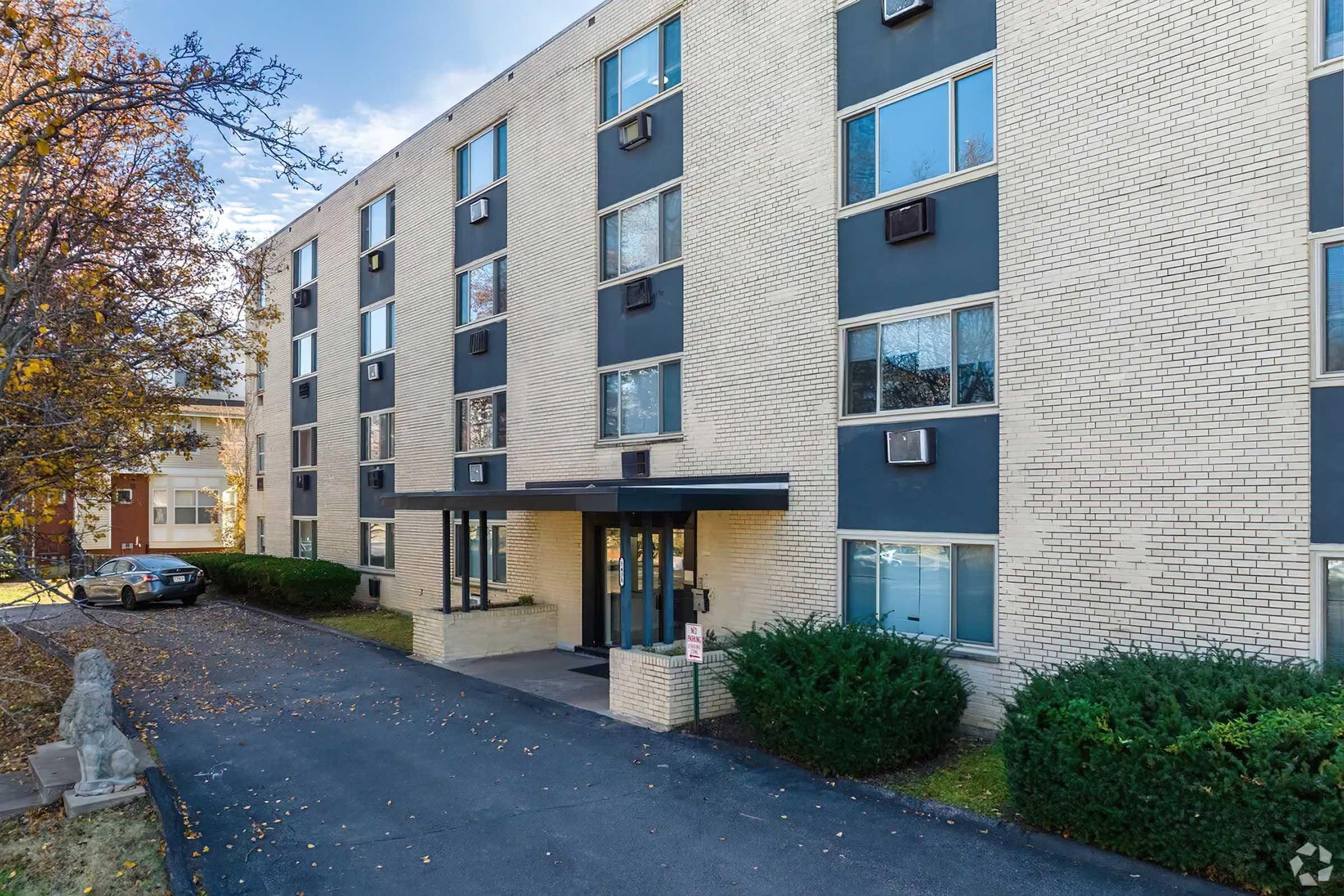 A view of a multi-story brick apartment building with large windows and several air conditioning units. The entrance is marked by a covered area with glass doors. Surrounding the building are manicured shrubs and a paved pathway, with a parked car visible in the background on a sunny day.