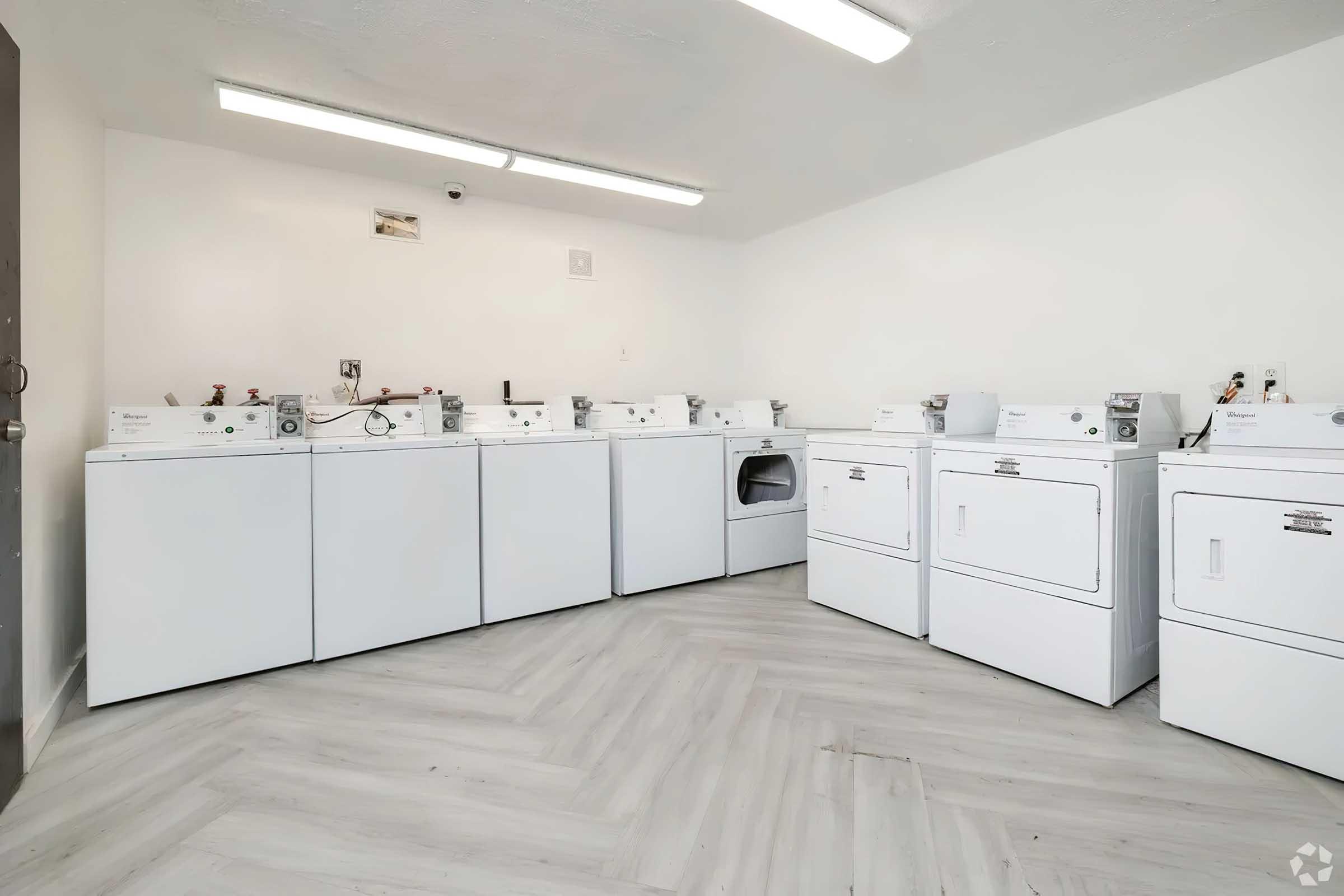 A clean, well-lit laundry room featuring multiple white washing machines and dryers arranged neatly against the walls, with wood-like flooring. The space has overhead lighting and a simple, uncluttered design, ideal for laundry tasks.