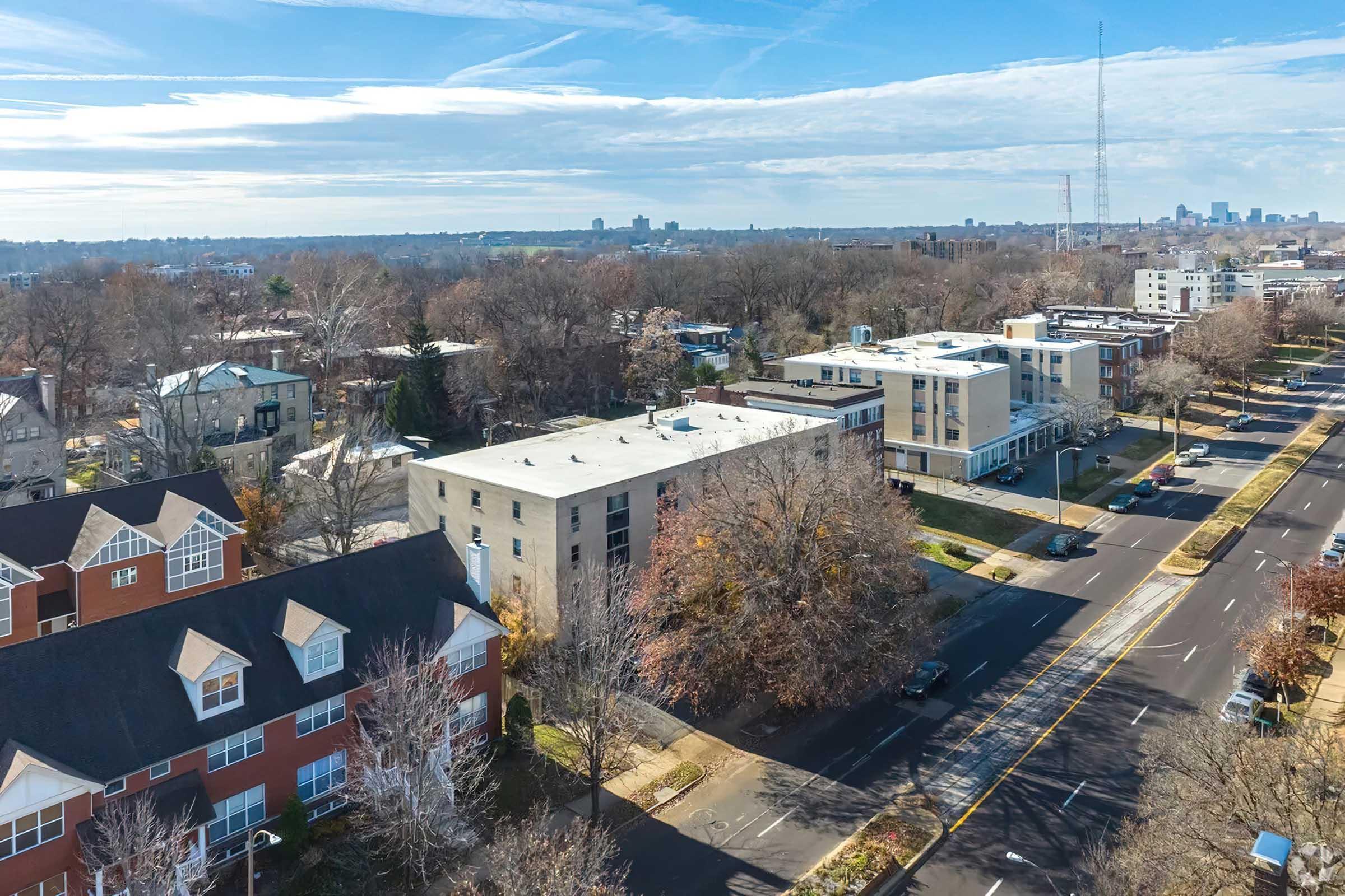Aerial view of a suburban area featuring residential buildings with a mix of architectural styles. Tree-lined streets and parked cars are visible, along with city skyline in the distance. The scene is set under a clear blue sky, indicating a sunny day.