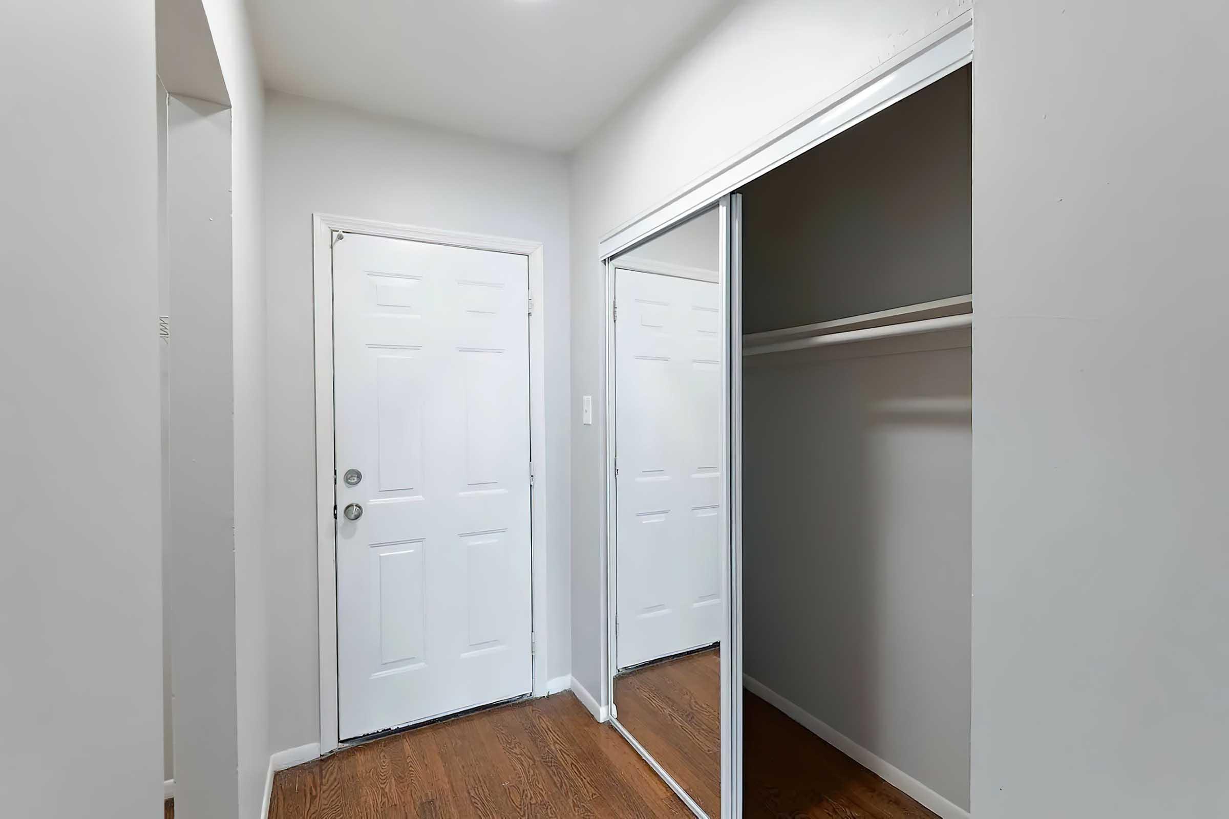 A hallway featuring a white interior with a door leading outside and a mirrored closet door on the right. The floor is wooden, and the walls are painted light gray. There is a small space near the door, creating a tidy and functional entryway.
