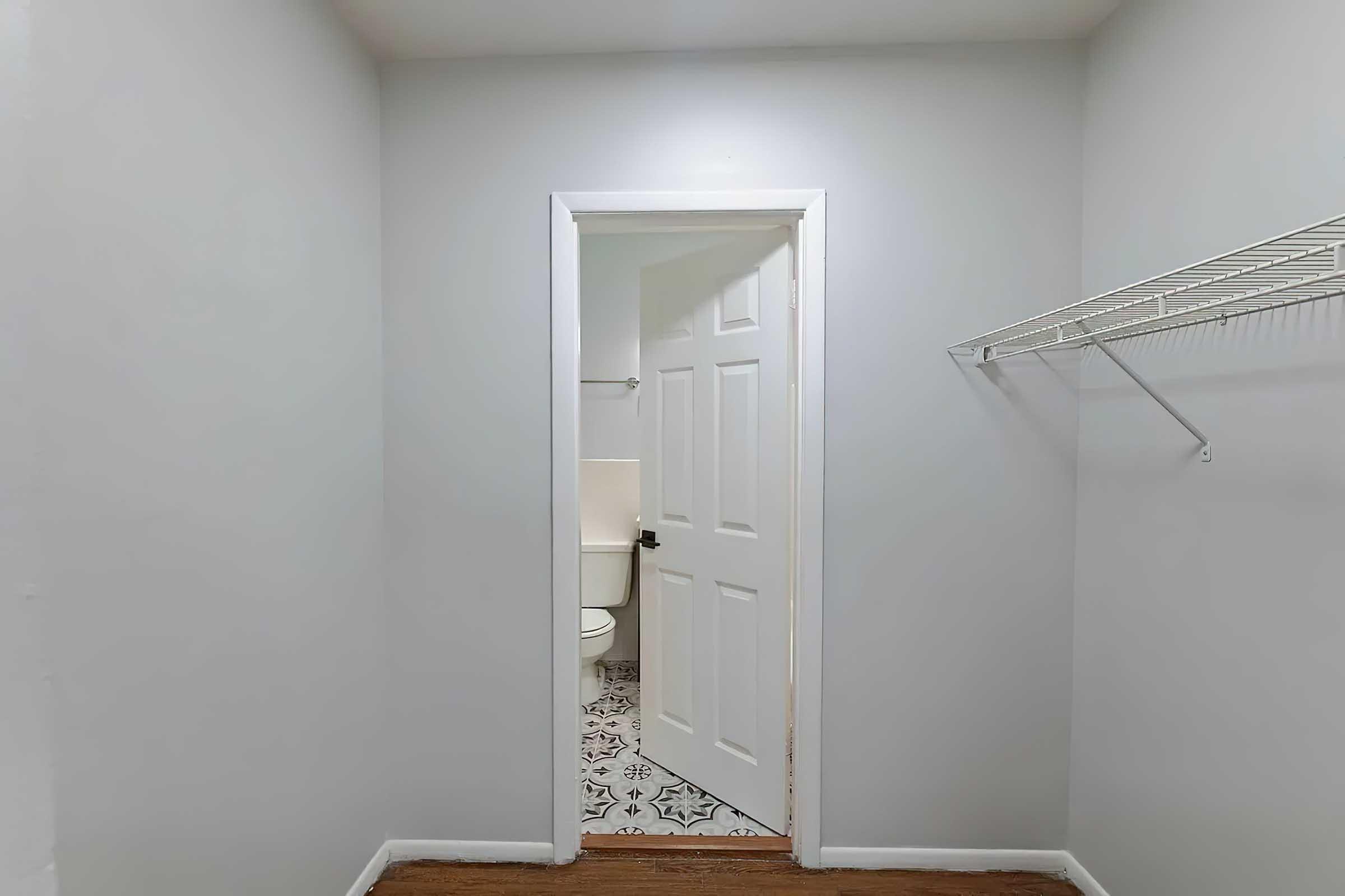 A small, empty room with light gray walls and hardwood flooring. A white door leads to a bathroom, visible in the background. A wire shelf is mounted on one wall, and there are no furnishings or decorations in the space.