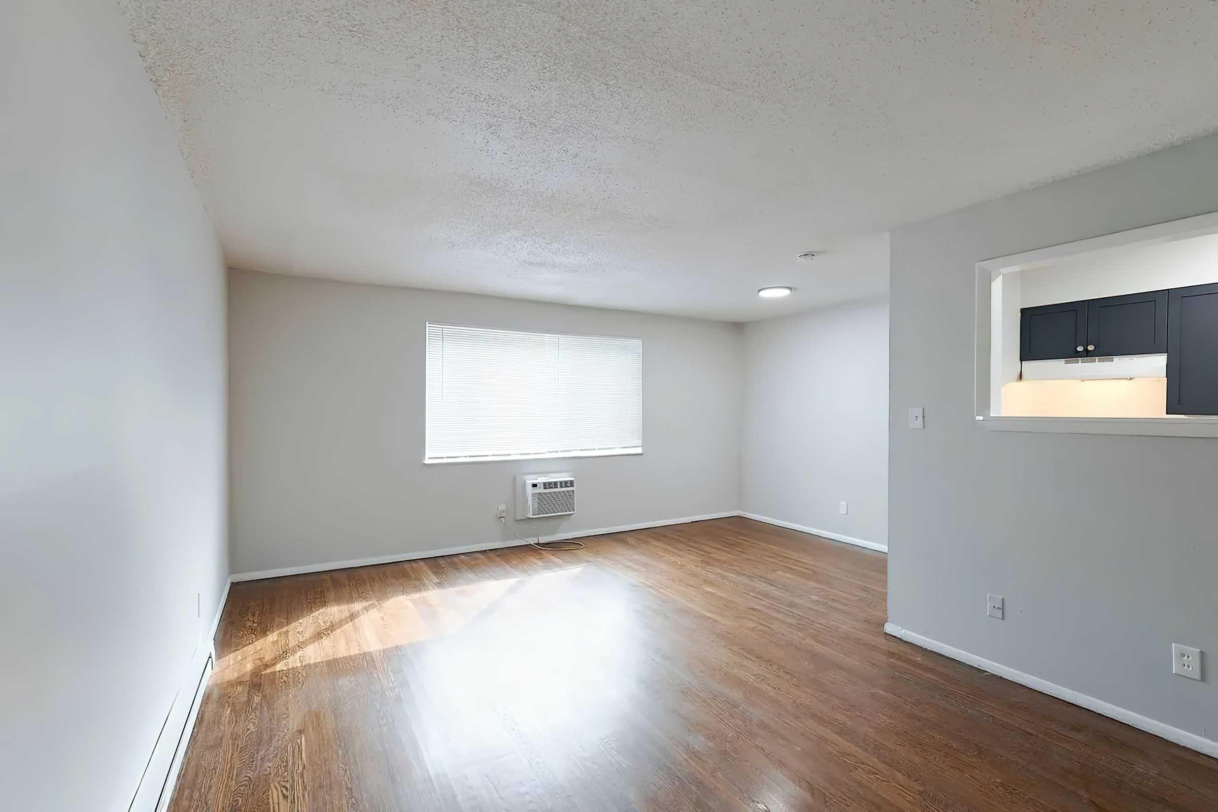 Spacious, bright room with hardwood flooring and a large window letting in natural light. An air conditioning unit is visible on the wall. The room features a small partition leading to a kitchenette area with dark cabinets. The walls are painted in neutral tones, creating a clean and modern atmosphere.