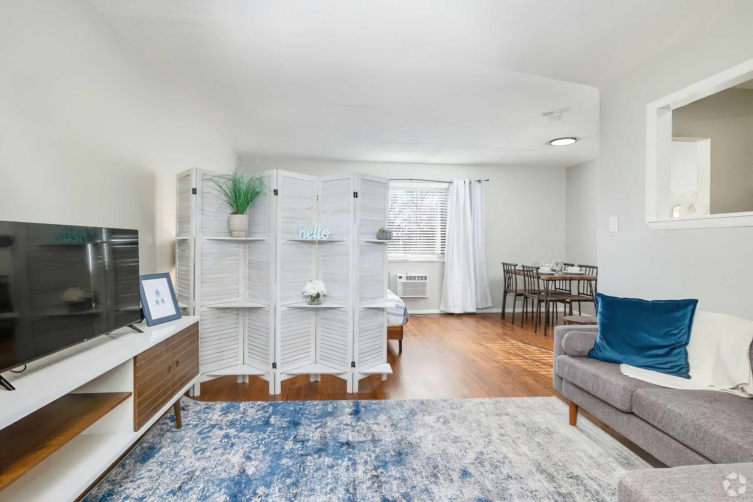 Bright living space featuring a comfortable gray sofa with blue accents, a wooden TV stand, and a decorative white screen. A dining area with a table and chairs is visible in the background, illuminated by natural light from a window with white curtains. The room has wooden flooring and a soft blue area rug.