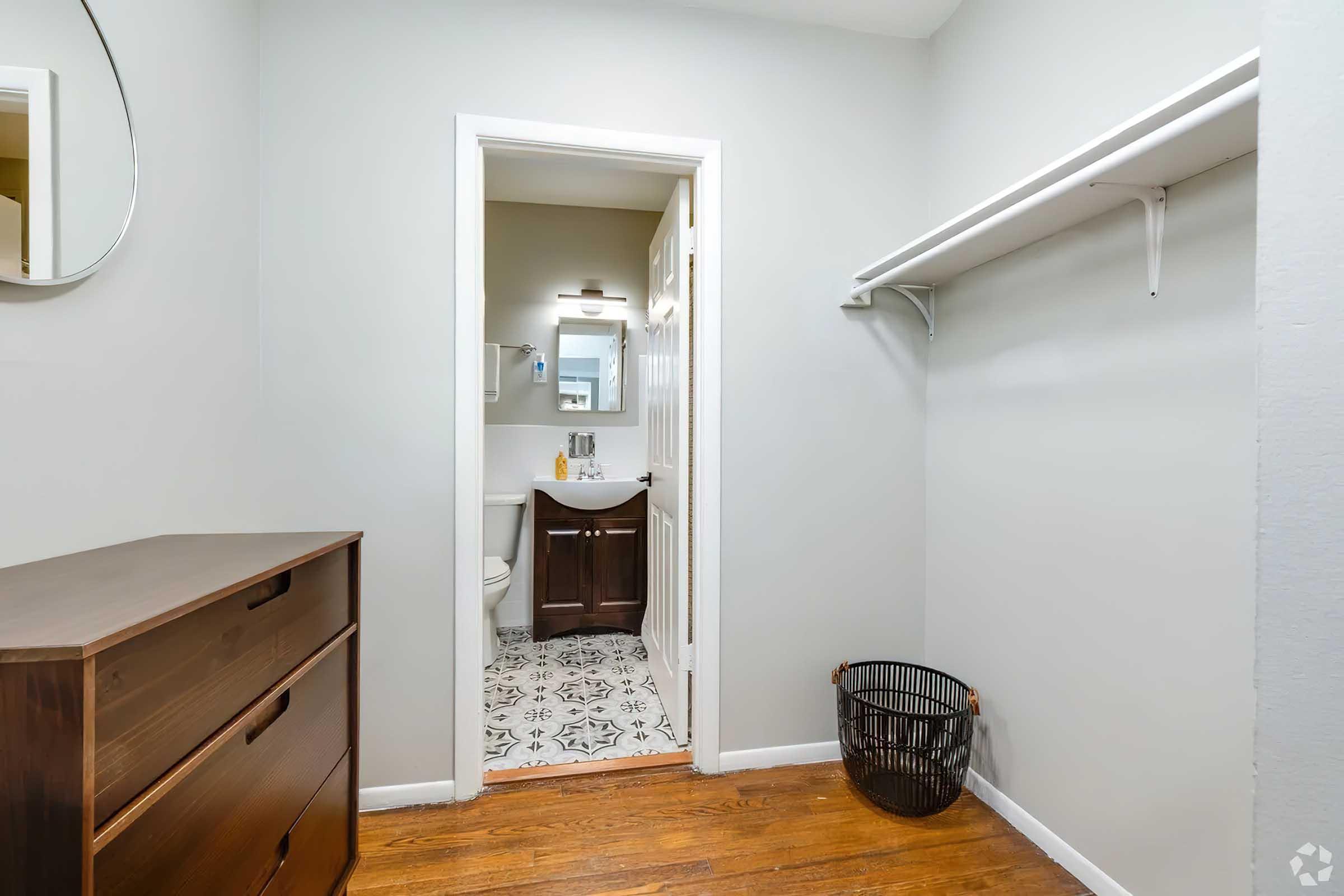 A small, well-lit room with gray walls. There is a wooden dresser on the left and a doorway leading to a bathroom. The bathroom features a mirror above a sink and modern fixtures. A black wire laundry basket is placed on the floor, and the flooring is wooden.