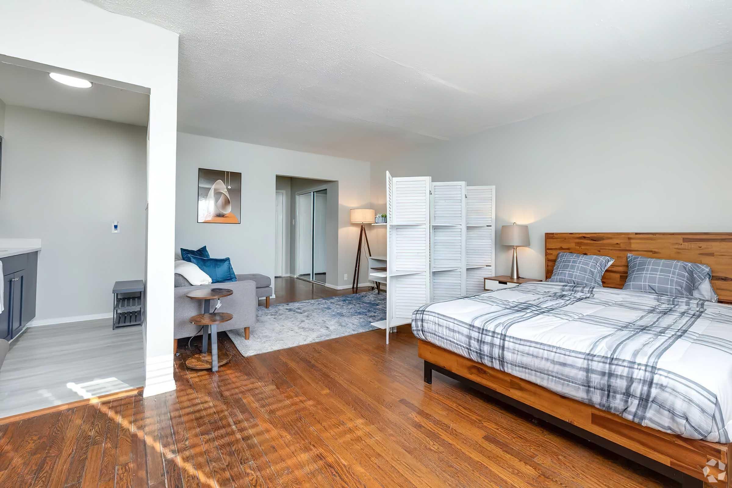 A well-lit bedroom featuring a wooden bed with a plaid comforter, a small side table, and a room divider. The space includes a plush gray sofa, a light blue area rug, and two lamps. There are doorways leading to other rooms, and the hardwood floor adds warmth to the modern decor.