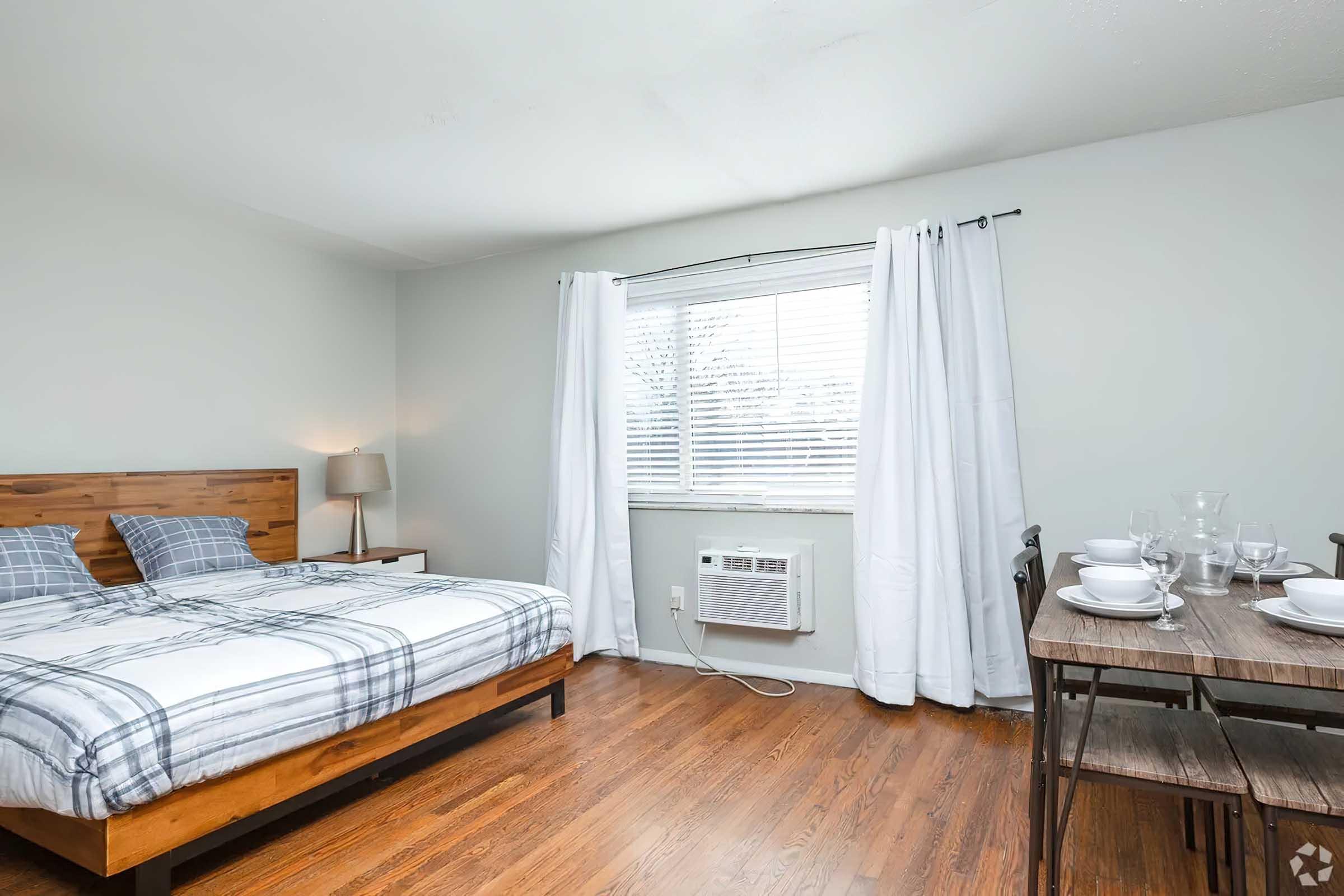 A cozy bedroom featuring a wooden bed with checkered bedding, a bedside lamp, and large white curtains. An air conditioning unit is visible under the window, and a small dining table set with plates and glasses is positioned nearby. The floor is made of hardwood, giving the room a warm ambiance.