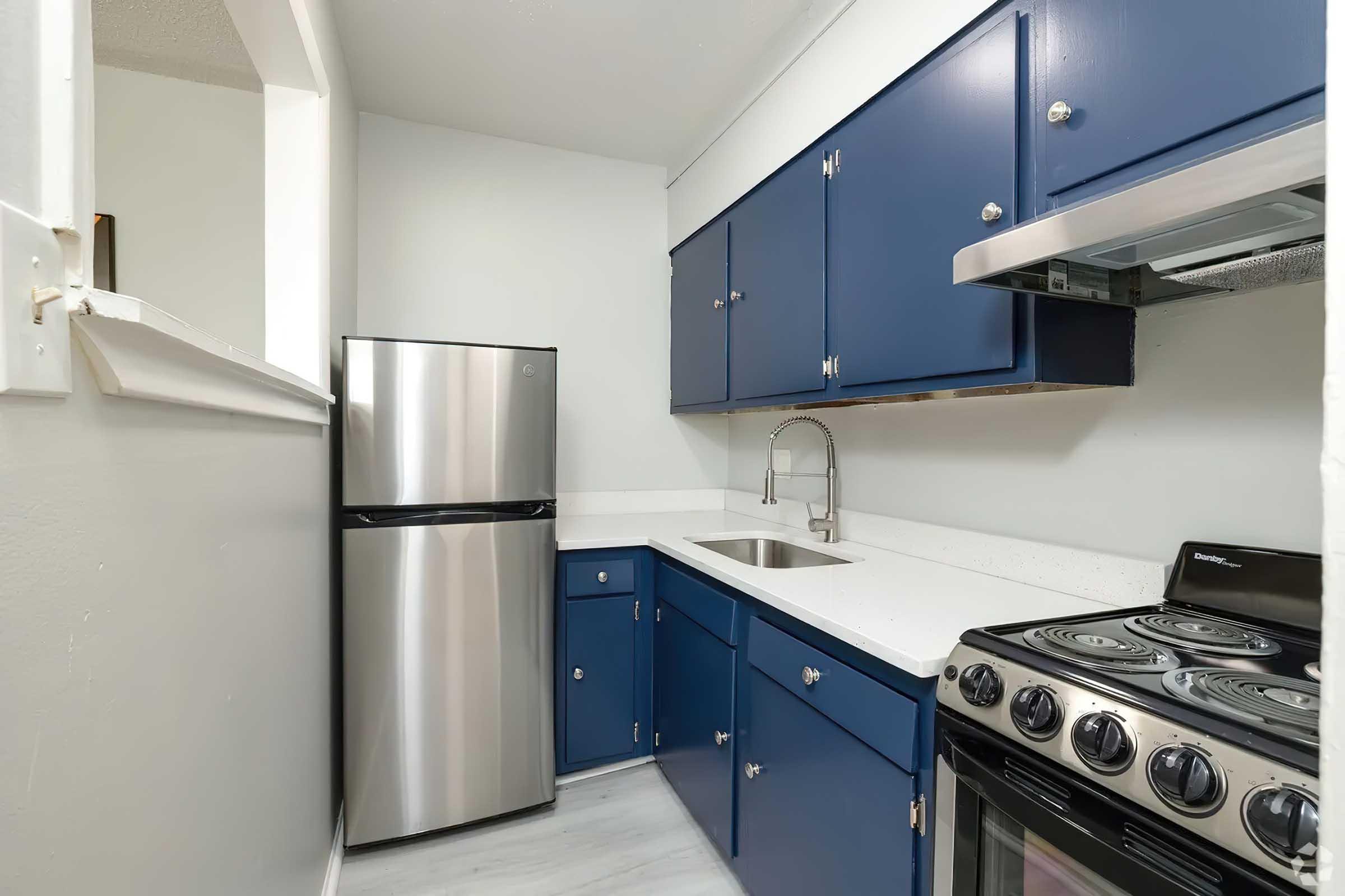 Compact kitchen featuring dark blue cabinets, a stainless steel refrigerator, a stainless steel sink, and a black gas stove. The countertops are white, and the kitchen has a clean, modern look with light-colored flooring and a neutral wall color.