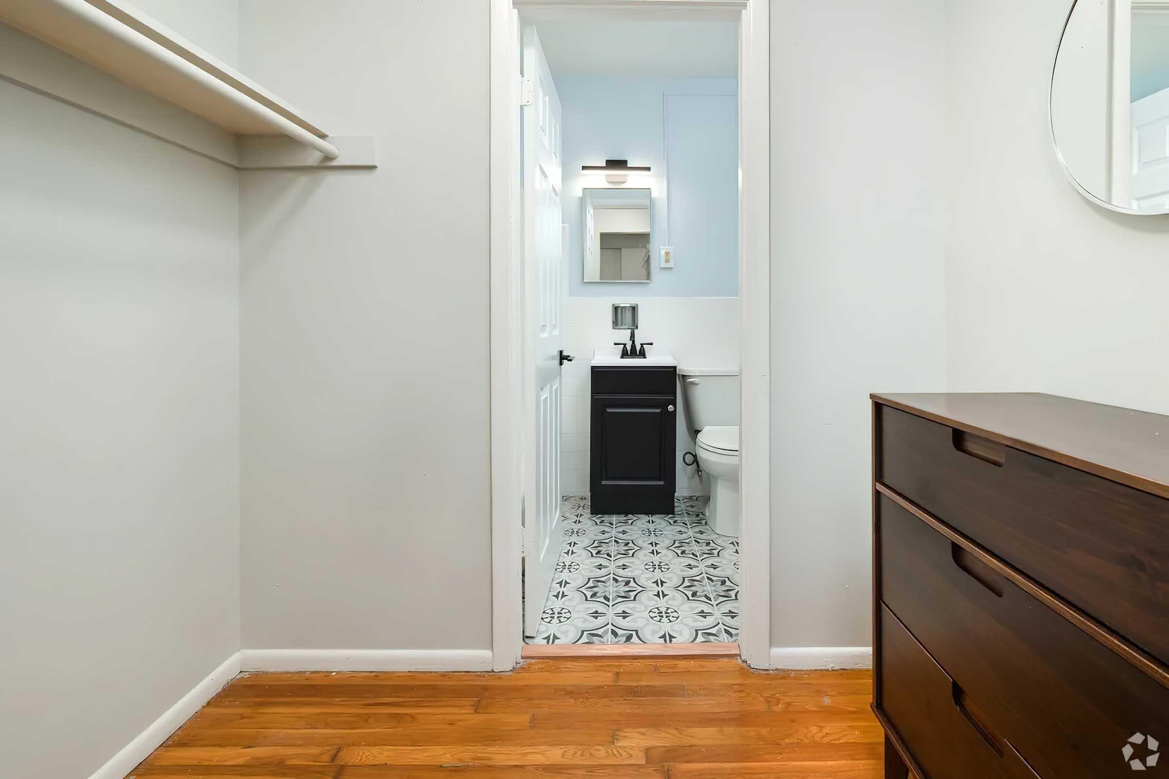 View of a small bathroom from a hallway. The image shows a doorway leading to a bathroom with a modern black vanity, a mirror, and a toilet. The floor features stylish patterned tiles, and there is a wooden dresser in the foreground with a simple design. The walls are painted in light colors, enhancing the space's brightness.