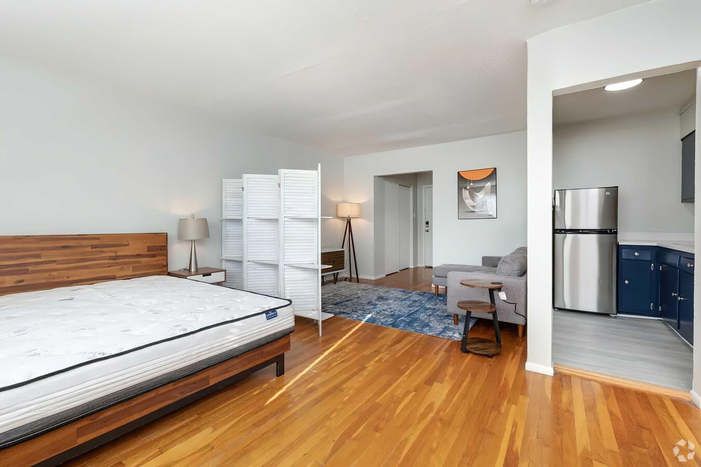 A spacious and well-lit bedroom featuring a wooden bed frame with a mattress, a room divider, two lamps, a small couch, and a blue area rug. The adjacent kitchenette is visible, showcasing a refrigerator and cabinets. The hardwood floor adds warmth to the modern decor.