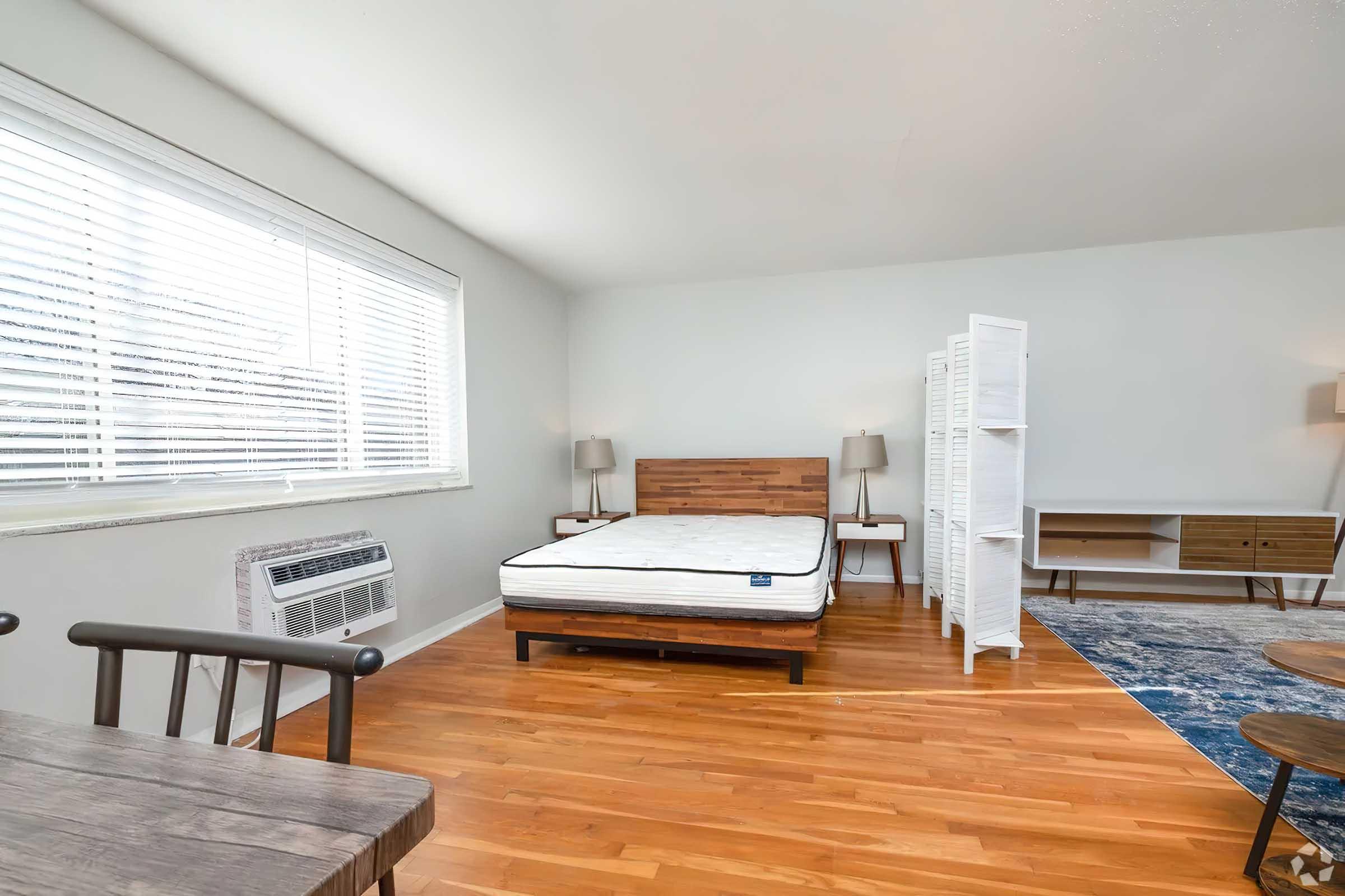 A spacious room featuring a wooden bed frame with a mattress, two lamps on nightstands, and a decorative room divider. The floor is hardwood, and there's a blue area rug near a wooden dining table with chairs. Large windows provide natural light, and an air conditioning unit is visible.