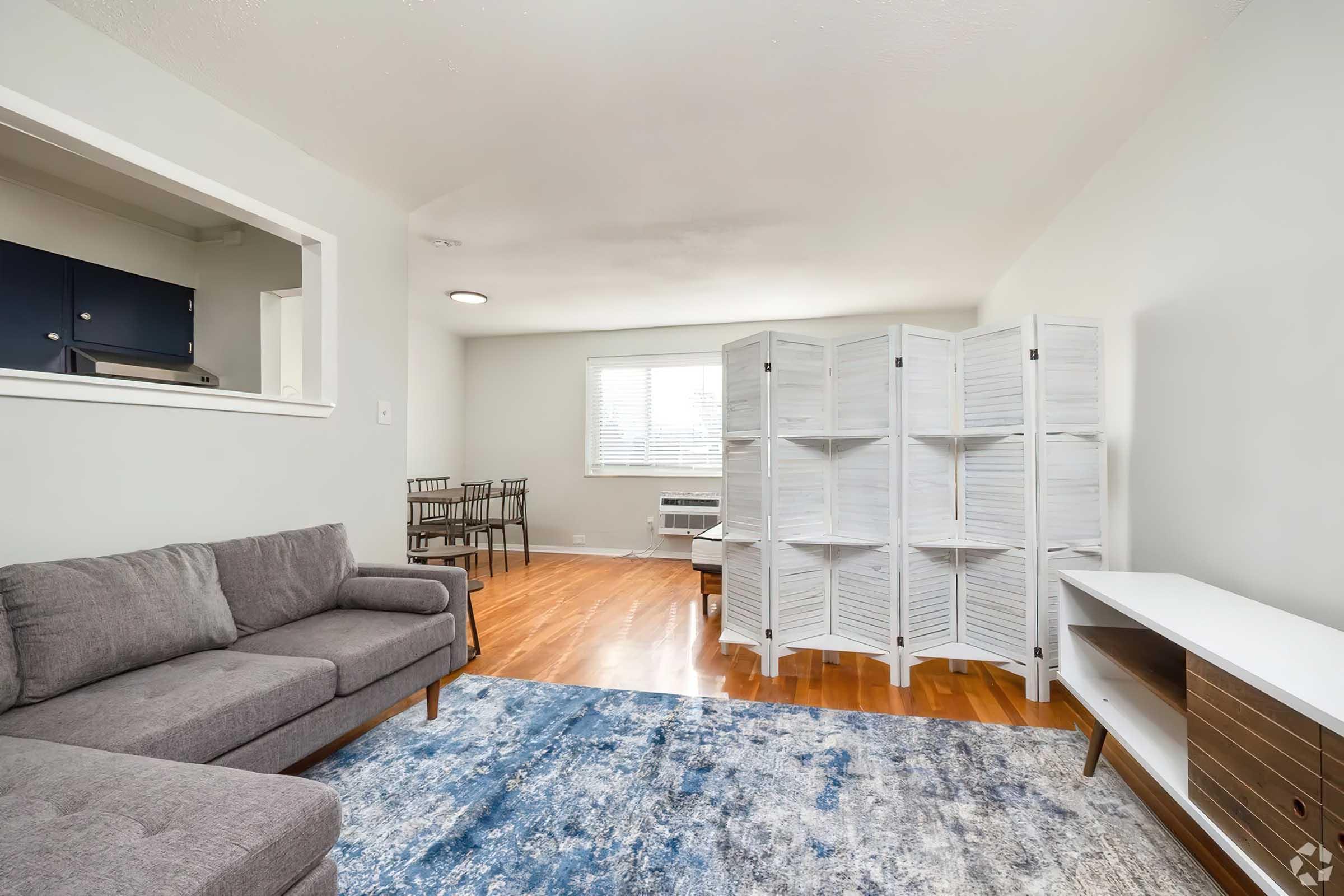 Cozy living room with a gray sofa, a white folding screen, and a wooden coffee table. A blue area rug covers the hardwood floor. There is a window with blinds allowing natural light, and a small dining area with chairs in the background. A media console is positioned against one wall.