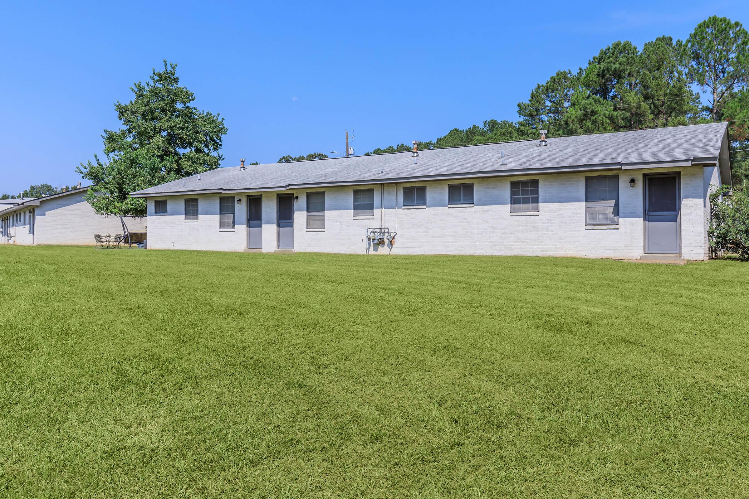 A view of a single-story residential building with multiple units, featuring light-colored brick walls, several windows, and a manicured green lawn. In the background, there are trees and a clear blue sky, creating a peaceful suburban atmosphere.