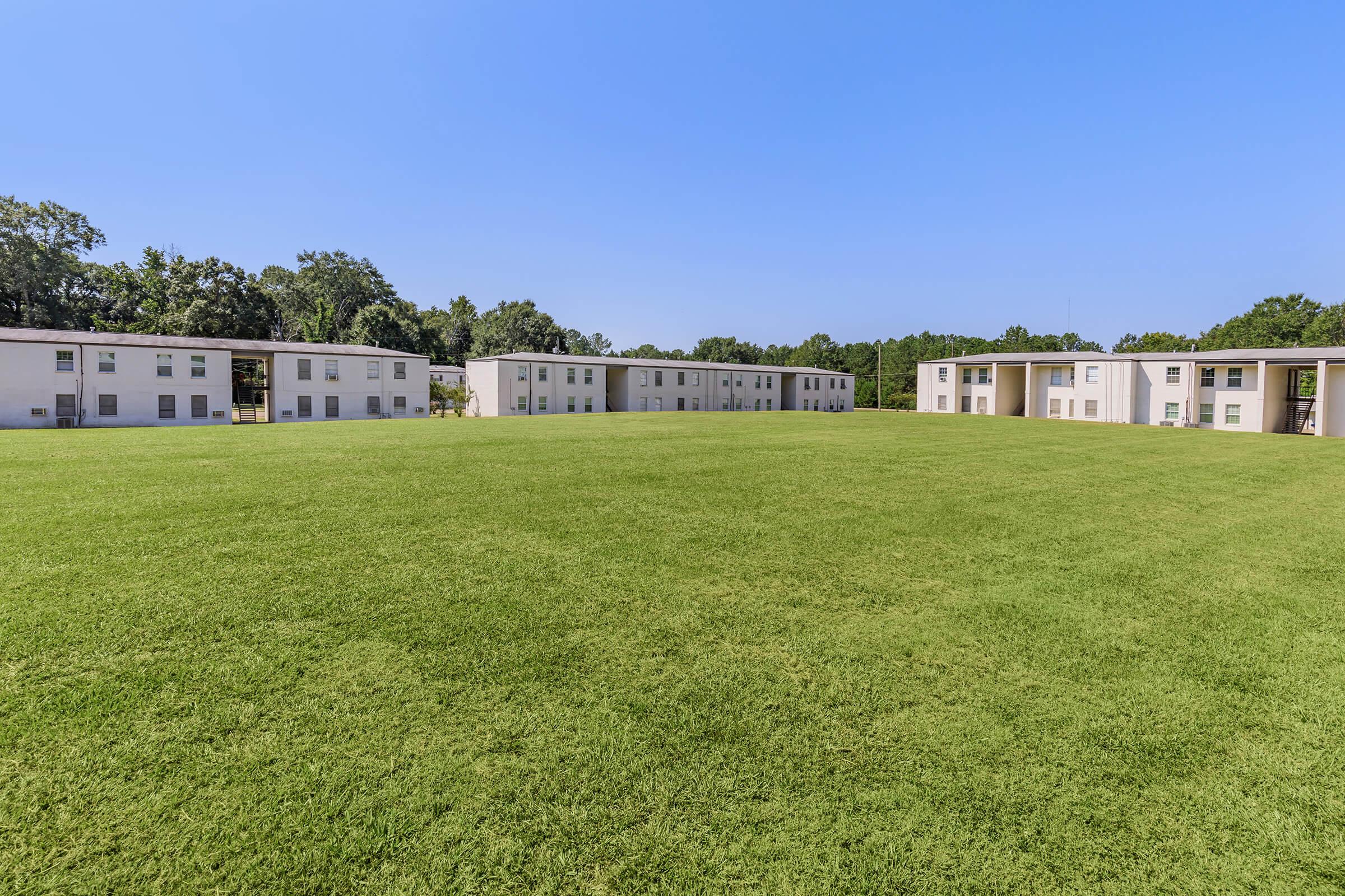Expansive green lawn surrounded by several white multi-unit residential buildings under a clear blue sky. The area features well-maintained grass and trees in the background, suggesting a peaceful suburban setting.