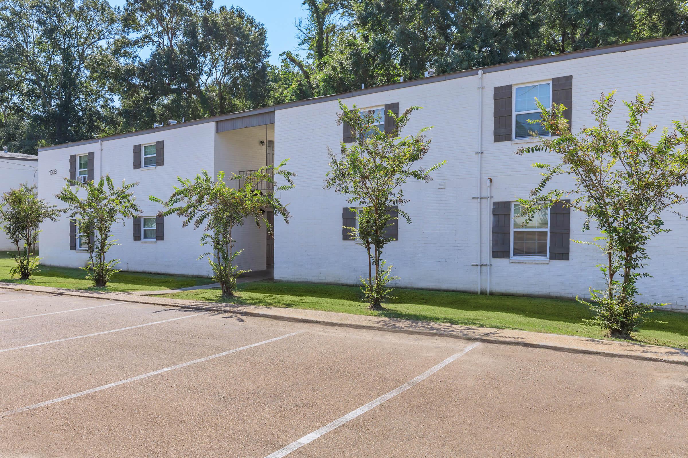 A white two-story apartment building with green landscaping in front. The exterior features multiple windows and a central entrance. There is a parking lot in the foreground with marked spaces, and trees line the walkway, providing a pleasant atmosphere.