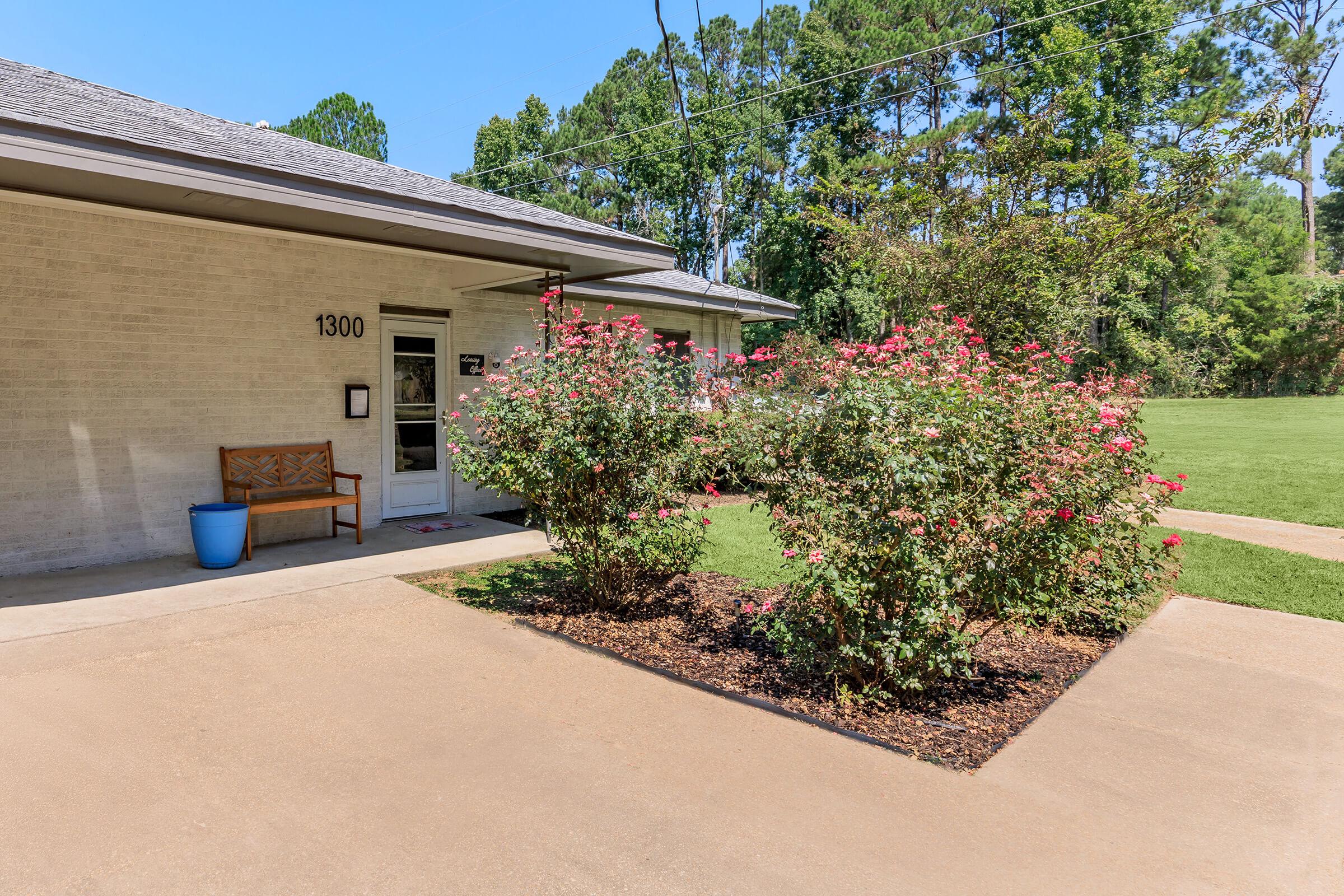 A sunny exterior view of a building with a numbered address (1300) visible. In front, there are blooming rose bushes with pink flowers, flanked by a simple wooden bench and a blue planter. Lush green grass and trees are in the background, creating a peaceful atmosphere.