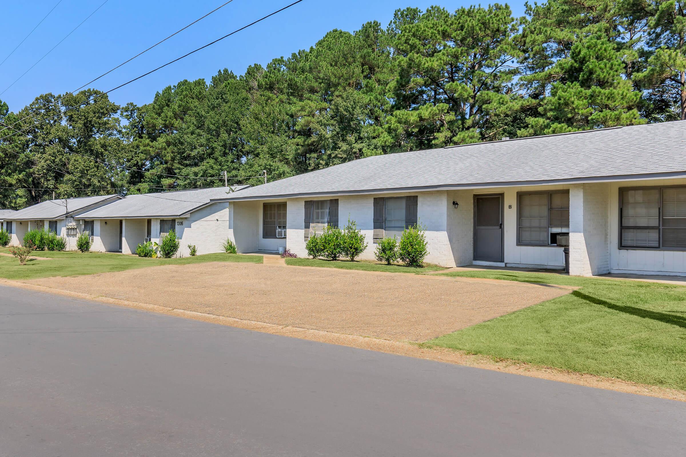 A row of single-story residential units with gray roofs, white siding, and large windows. The well-maintained lawn features neatly trimmed grass and small shrubs. In the background, dense trees provide a natural setting, and a paved road runs alongside the buildings. Clear blue sky above.