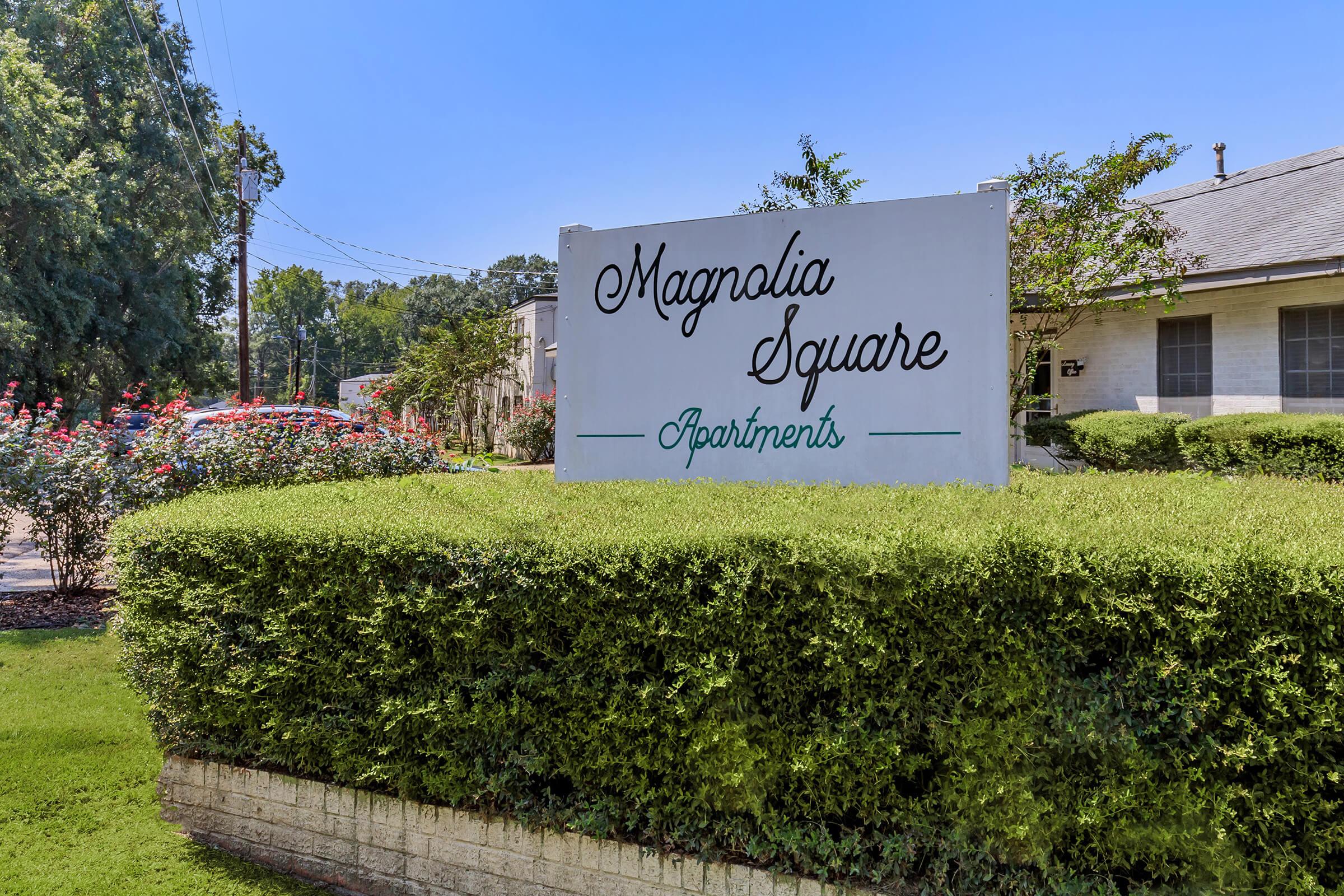 Sign for Magnolia Square Apartments, surrounded by green shrubs and colorful flowers, with clear blue sky in the background. The sign features decorative lettering and is positioned at the entrance of the apartment complex.