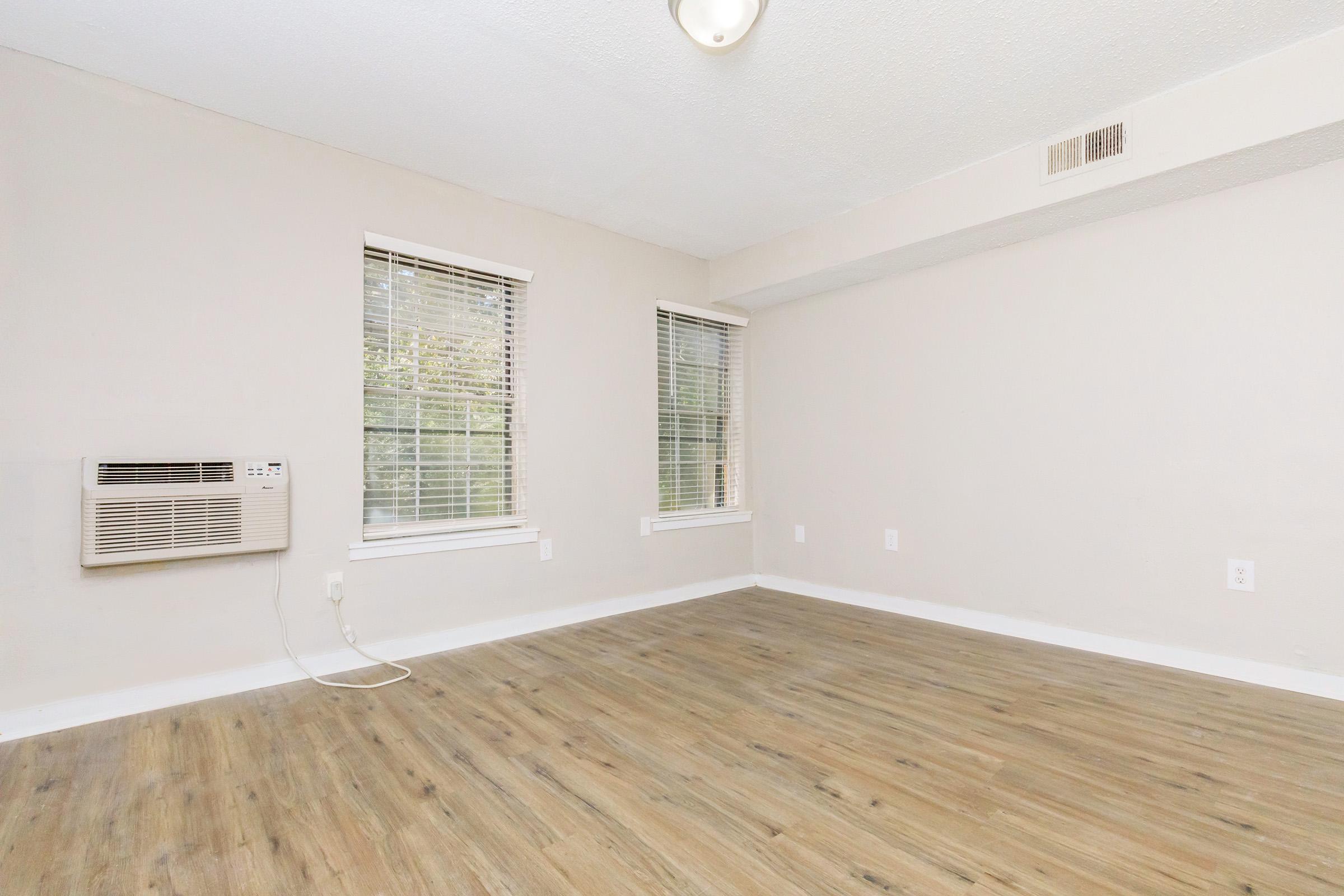 Empty room with light-colored walls and wooden flooring. Two windows with blinds allow natural light. An air conditioning unit is mounted on the wall. The space is uncluttered, creating a bright and airy atmosphere.
