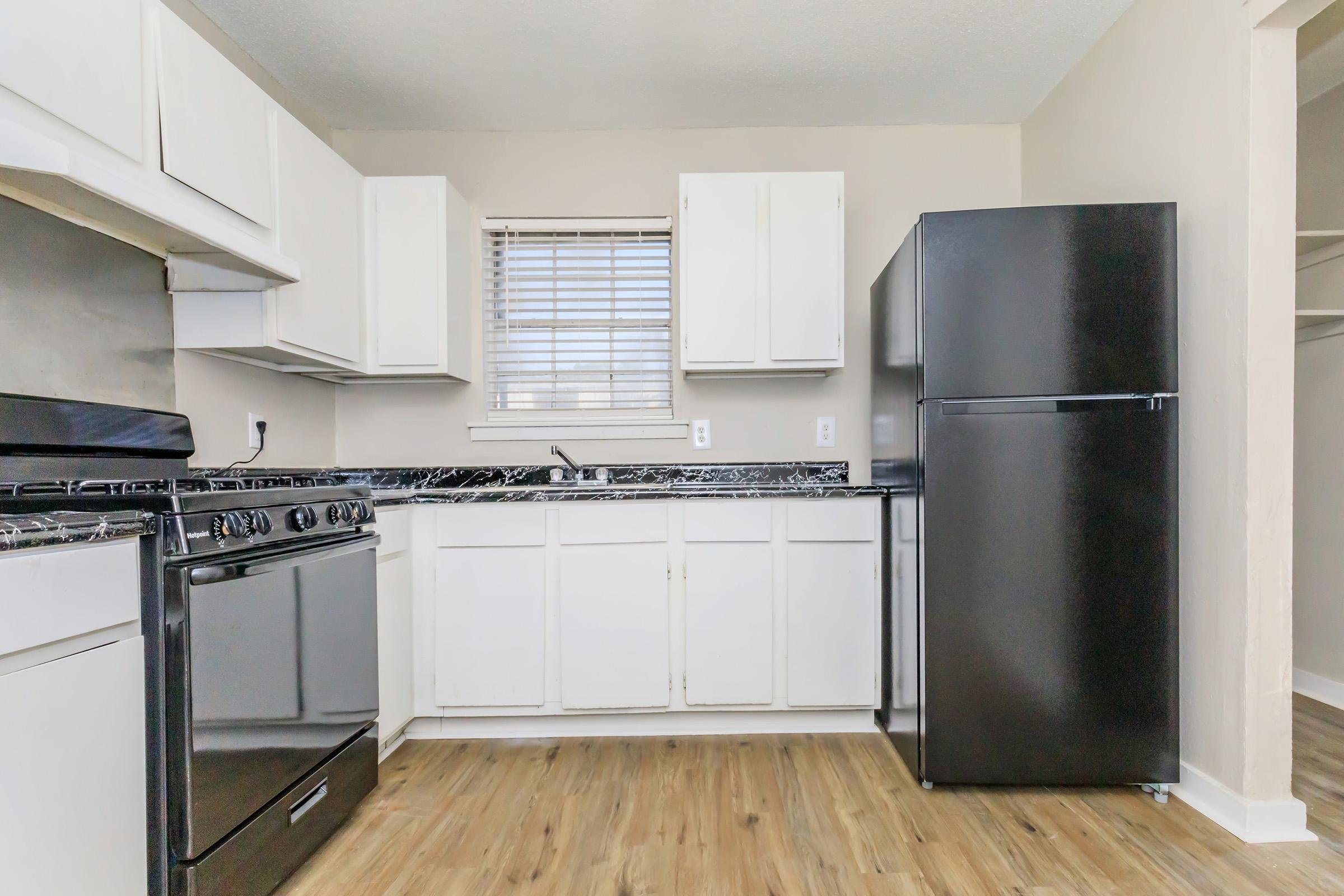A modern kitchen featuring white cabinets, a black refrigerator, and a black gas stove. The countertop has a dark, marbled design. A window provides natural light, and the floor is made of light wood planks.