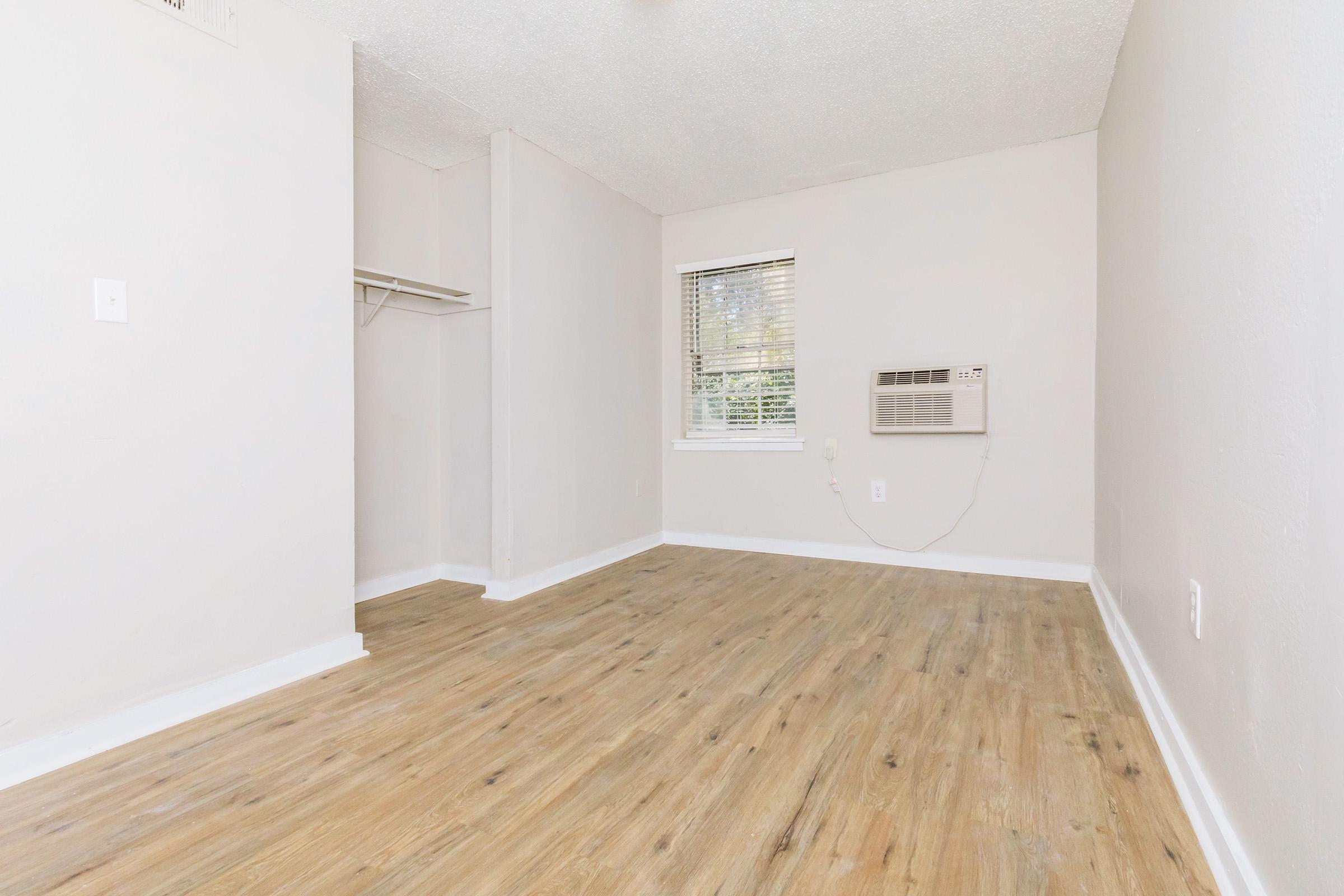 A small, empty room with light-colored walls and wood-like flooring. There is a window with blinds, an air conditioning unit mounted on the wall, and a small closet on the left side. The room is well-lit, creating a spacious and airy feel.