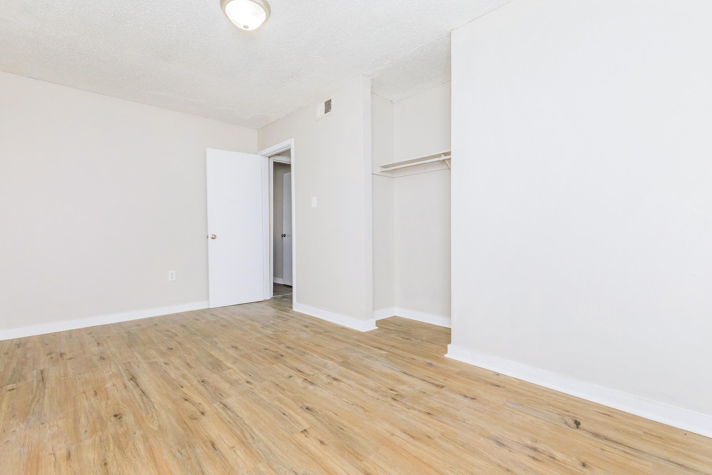 Empty room with light-colored walls and wooden flooring. A door is partially visible on the left, leading to another space. There is a closet with a single shelf on the right side, and a ceiling light fixture. Natural light brightens the room, creating a spacious and clean appearance.