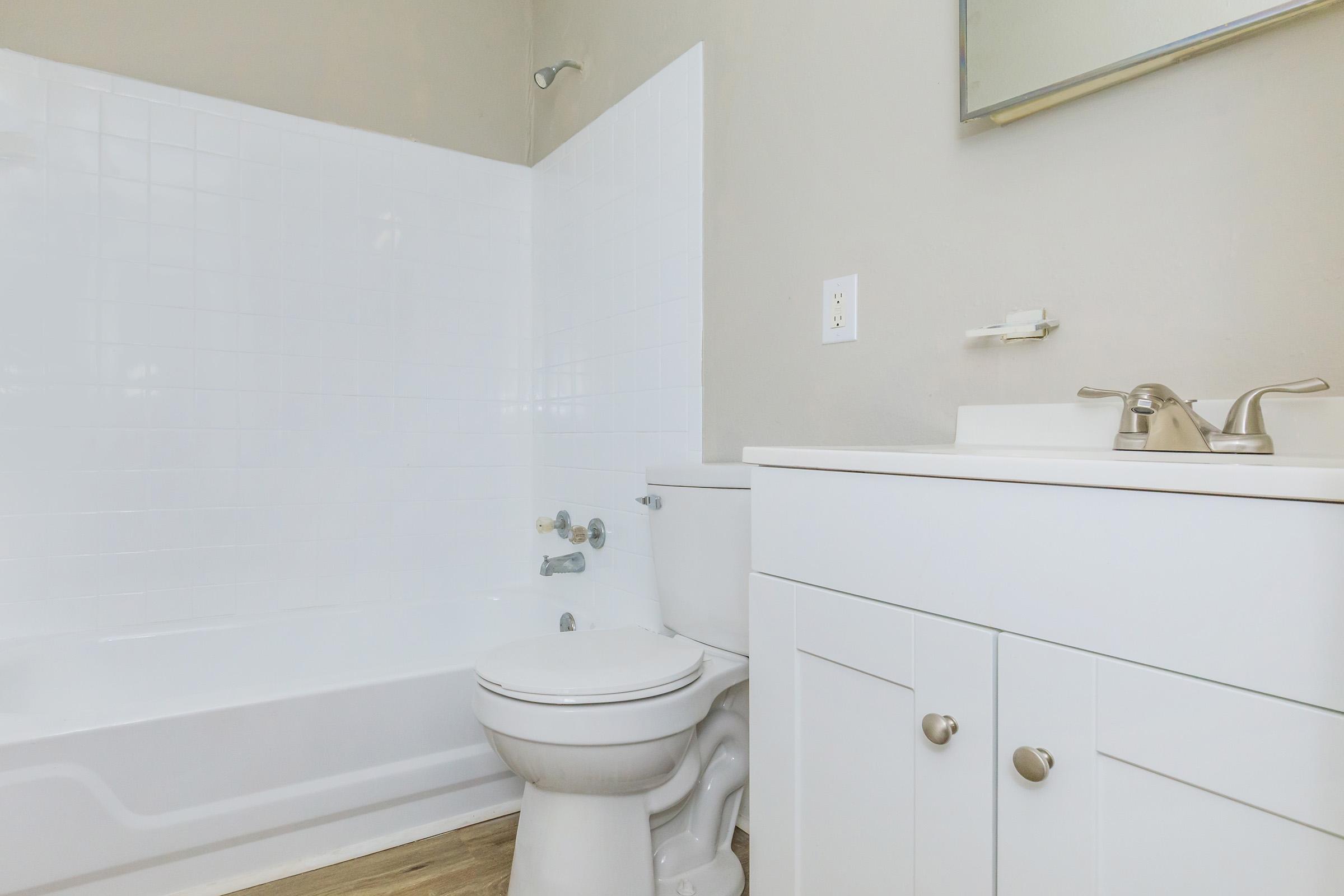 Clean and modern bathroom featuring a white bathtub, toilet, and sink with a cabinet. The walls are painted a light color, and there is a large mirror above the sink. The flooring is light wood, enhancing the contemporary design of the space.
