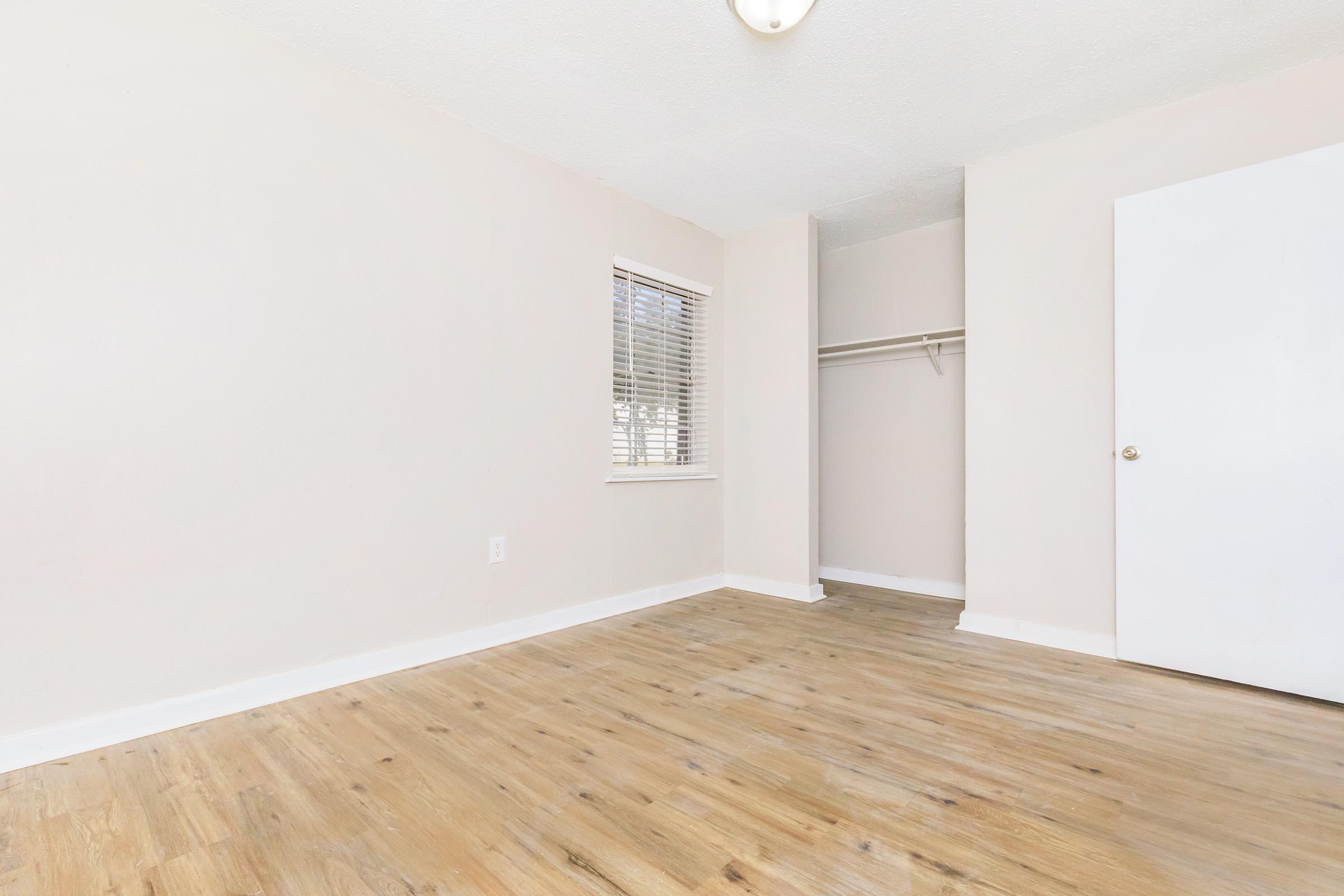 Empty room with light-colored walls and wooden flooring. A window with blinds allows natural light to enter. There is a small closet on the left side of the room, featuring a shelf and hanging space. The door on the right leads to another area, contributing to a clean and spacious atmosphere.