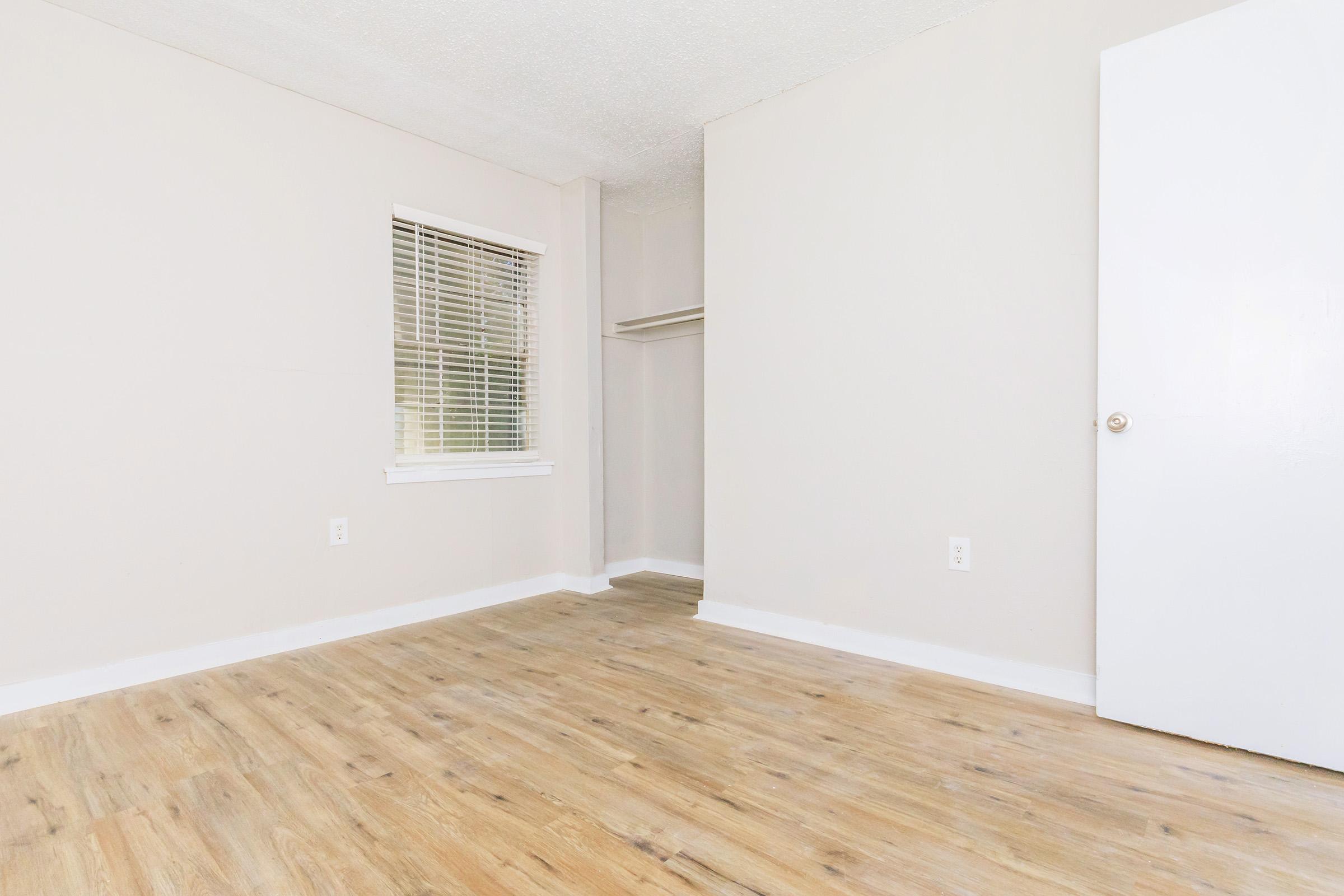 A vacant room with light-colored walls and a wooden floor. It features a window with blinds on one side and a closet space in the corner. The doorway on the right shows a simple white door. The room is well-lit and has a clean, minimalist appearance.