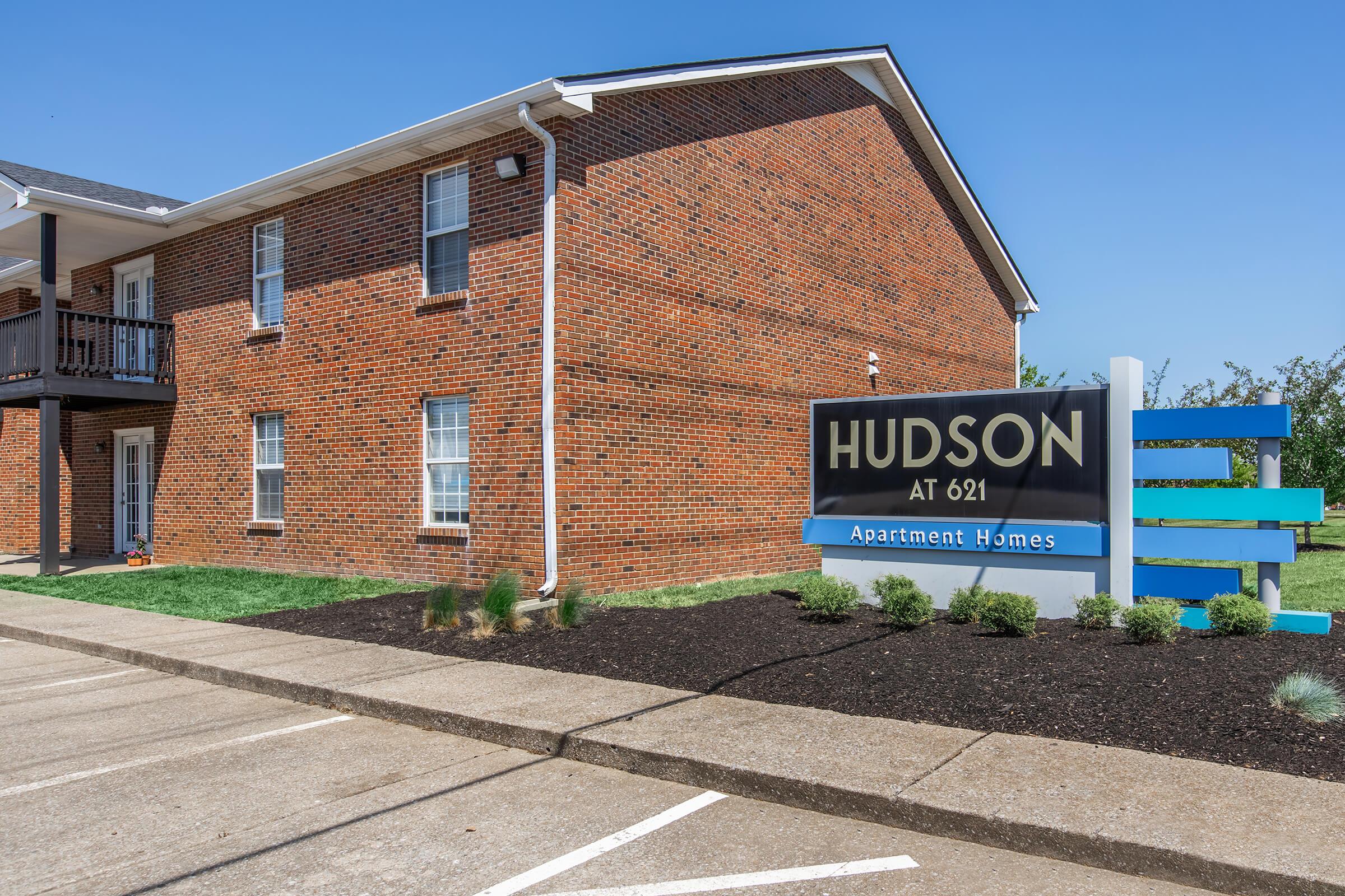 Brick building with multiple apartment units, featuring a prominent sign that reads "HUDSON AT 621 Apartment Homes." The landscape includes well-manicured grass and a clear blue sky. Parking spaces are visible in front of the building.
