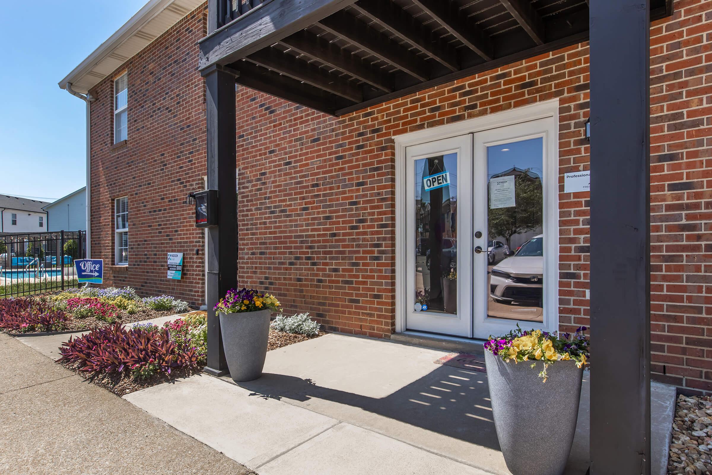 Entrance to a brick building featuring two large potted plants with colorful flowers on either side of a concrete walkway. The glass double doors display an "OPEN" sign. Visible in the background is a swimming pool area, and there are well-maintained shrubs and landscaping near the entrance.