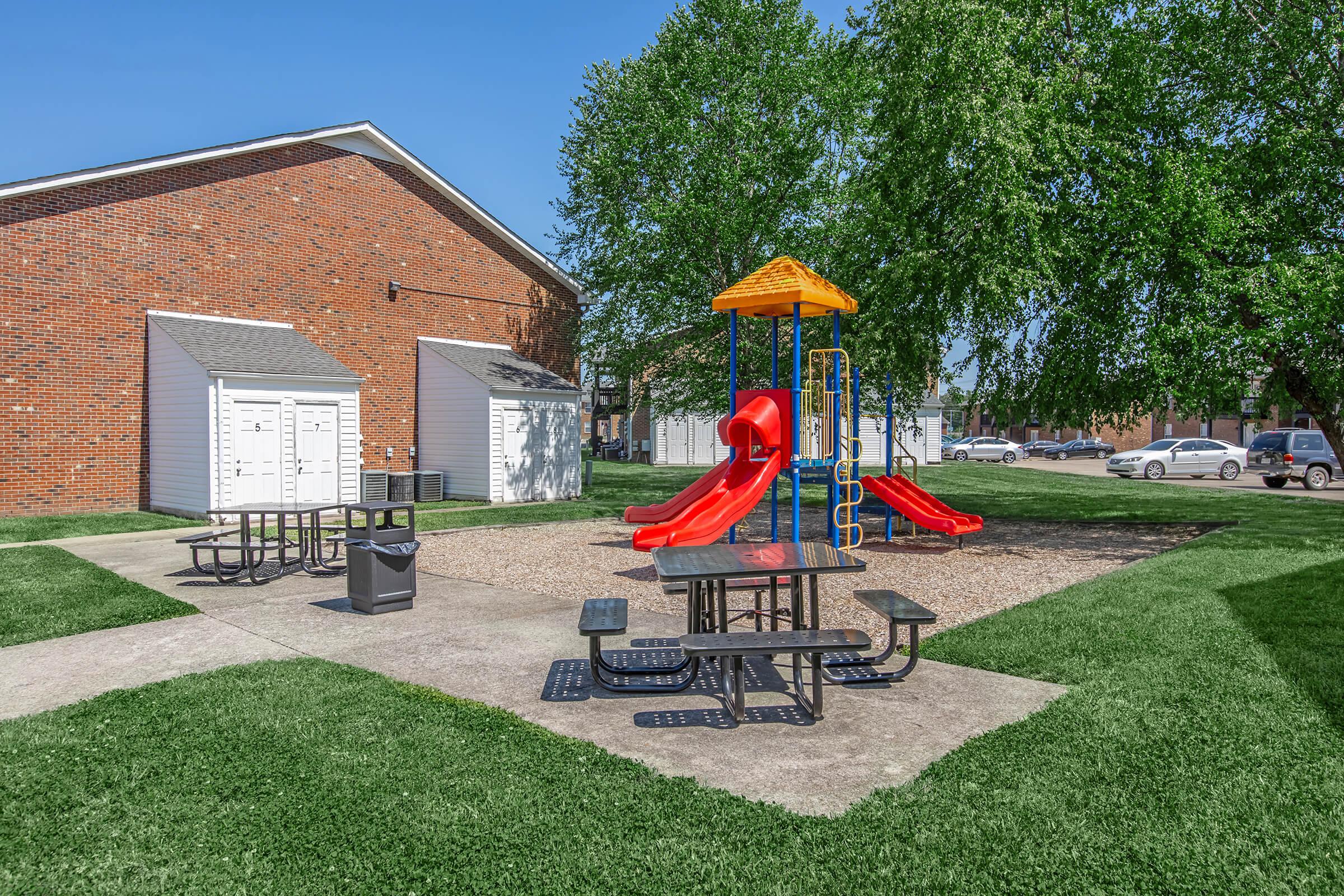 Playground area featuring a red and yellow slide structure surrounded by gravel, with picnic tables and a trash bin nearby. Green grass and trees create a pleasant outdoor environment, and a brick building is visible in the background. Clear blue sky above adds to the cheerful scene.