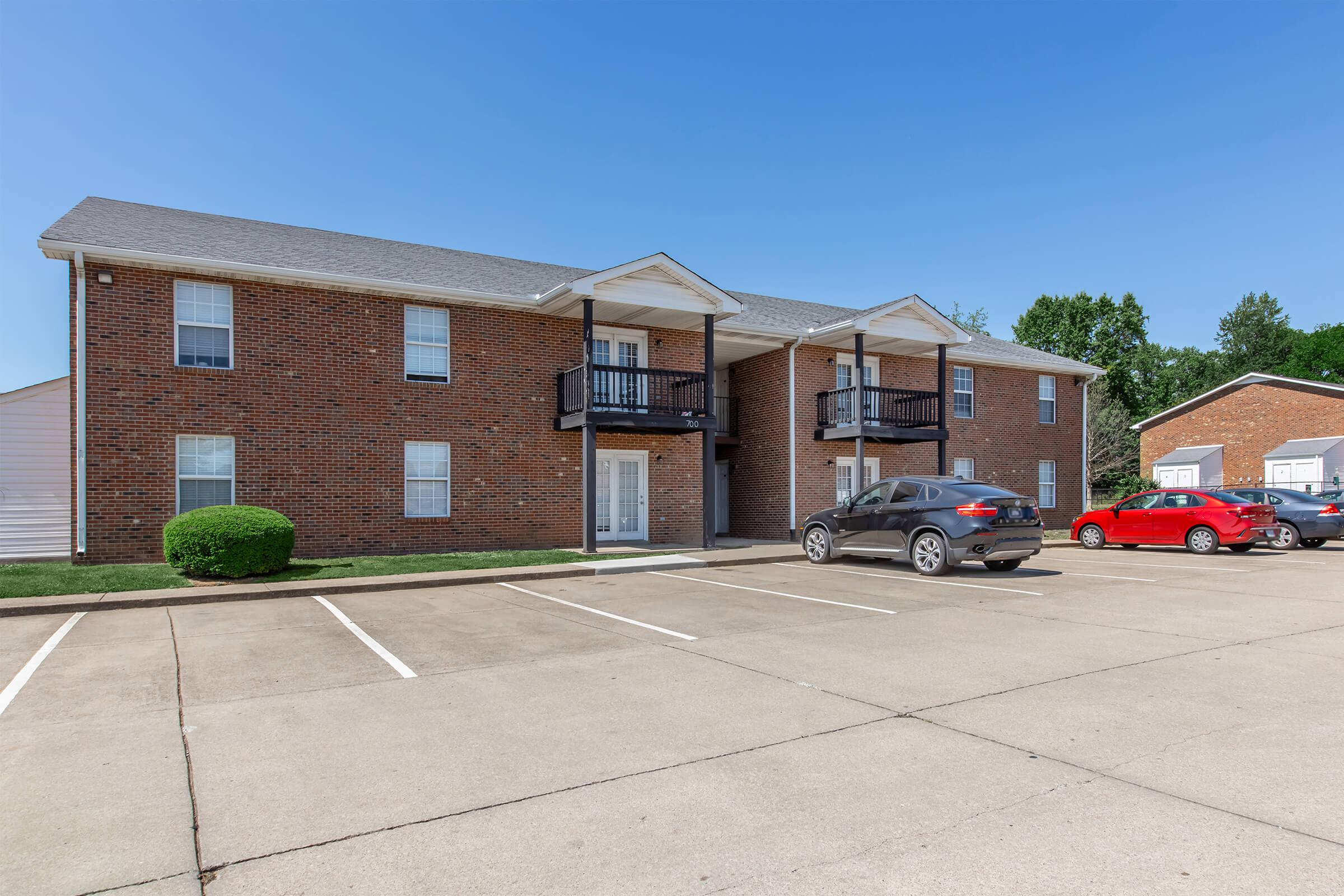 A brick apartment building with two levels and balconies, situated in a suburban area. There is a parking lot in front with several cars parked, and a grassy area along the building's foundation. Clear blue sky overhead.