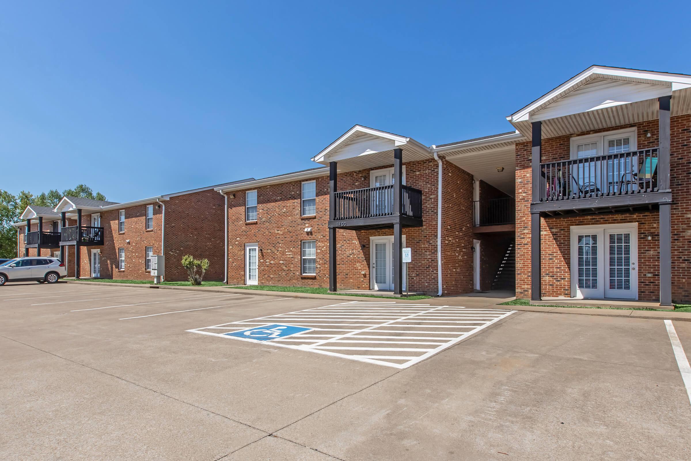 A view of a brick apartment complex featuring multiple two-story buildings. Each building has balconies and stairs leading to the upper units. A designated handicap parking space is visible in the foreground, along with several open parking spots. The sky is clear and blue, indicating a sunny day.