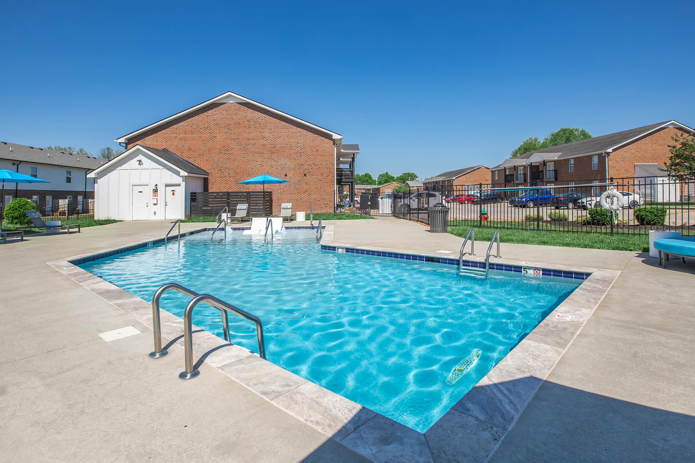 A clear blue swimming pool surrounded by a concrete deck. Two lounge chairs with umbrellas are near the pool, and a few apartment buildings are visible in the background. The sky is bright and sunny, creating a cheerful outdoor atmosphere.